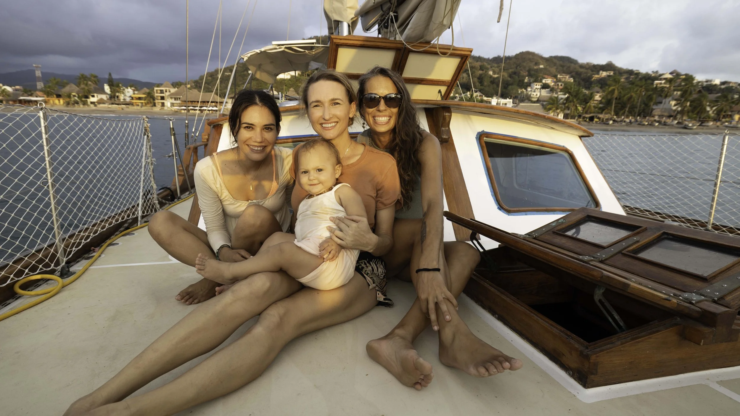 Four women and a young girl sitting together on the deck of a boat, with a scenic harbor and hillside town in the background.