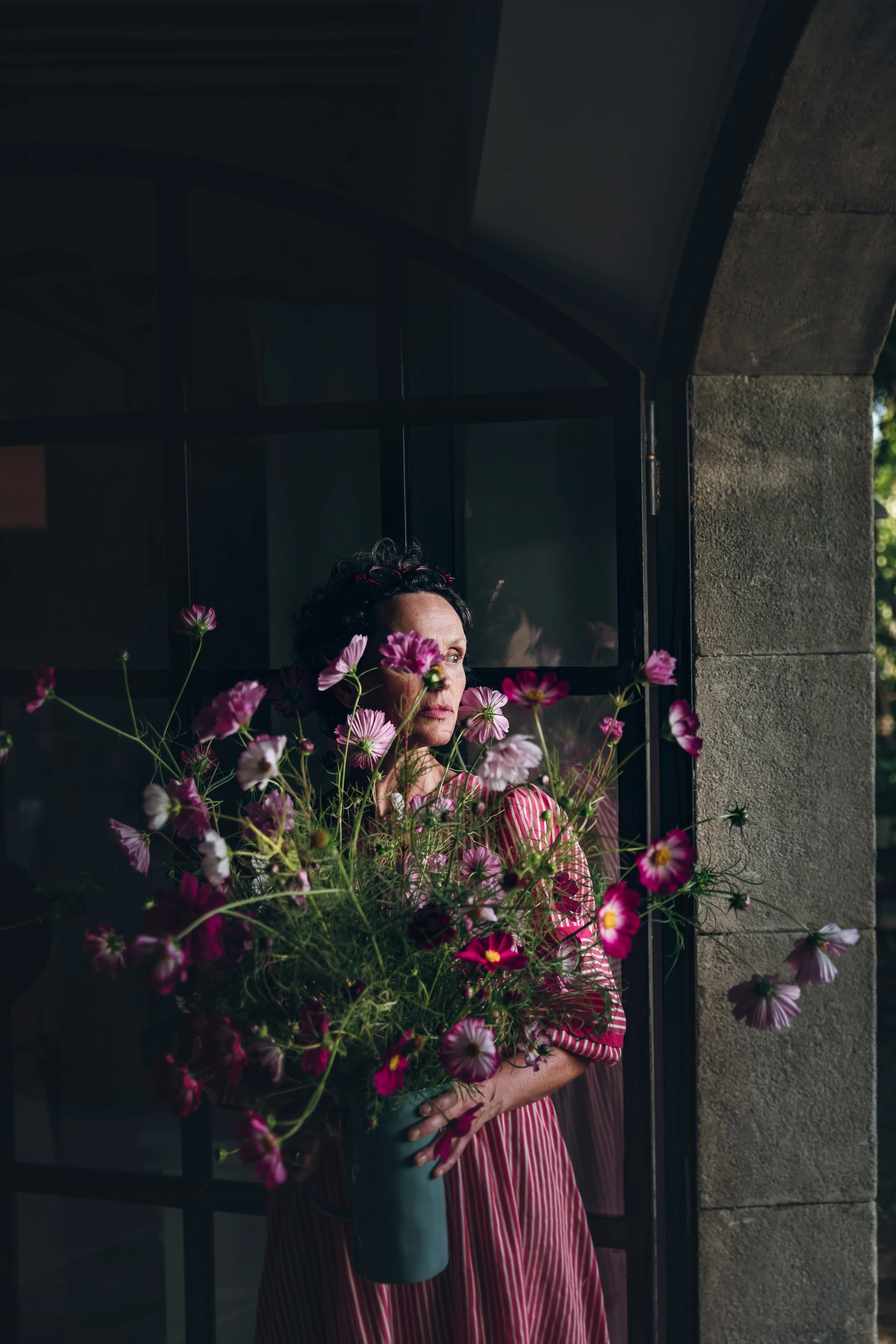 A woman standing by a window, holding a large bouquet of pink and purple flowers, looking out thoughtfully.