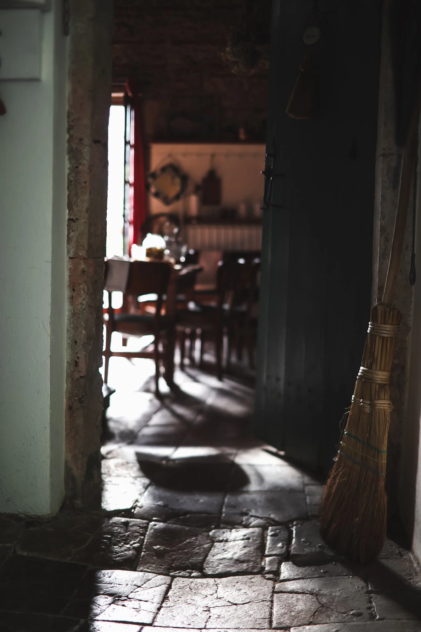 View through a doorway into a rustic dining room, with a broom leaning against the wall on the right, sunlight streaming in from a window and casting shadows on a stone floor.