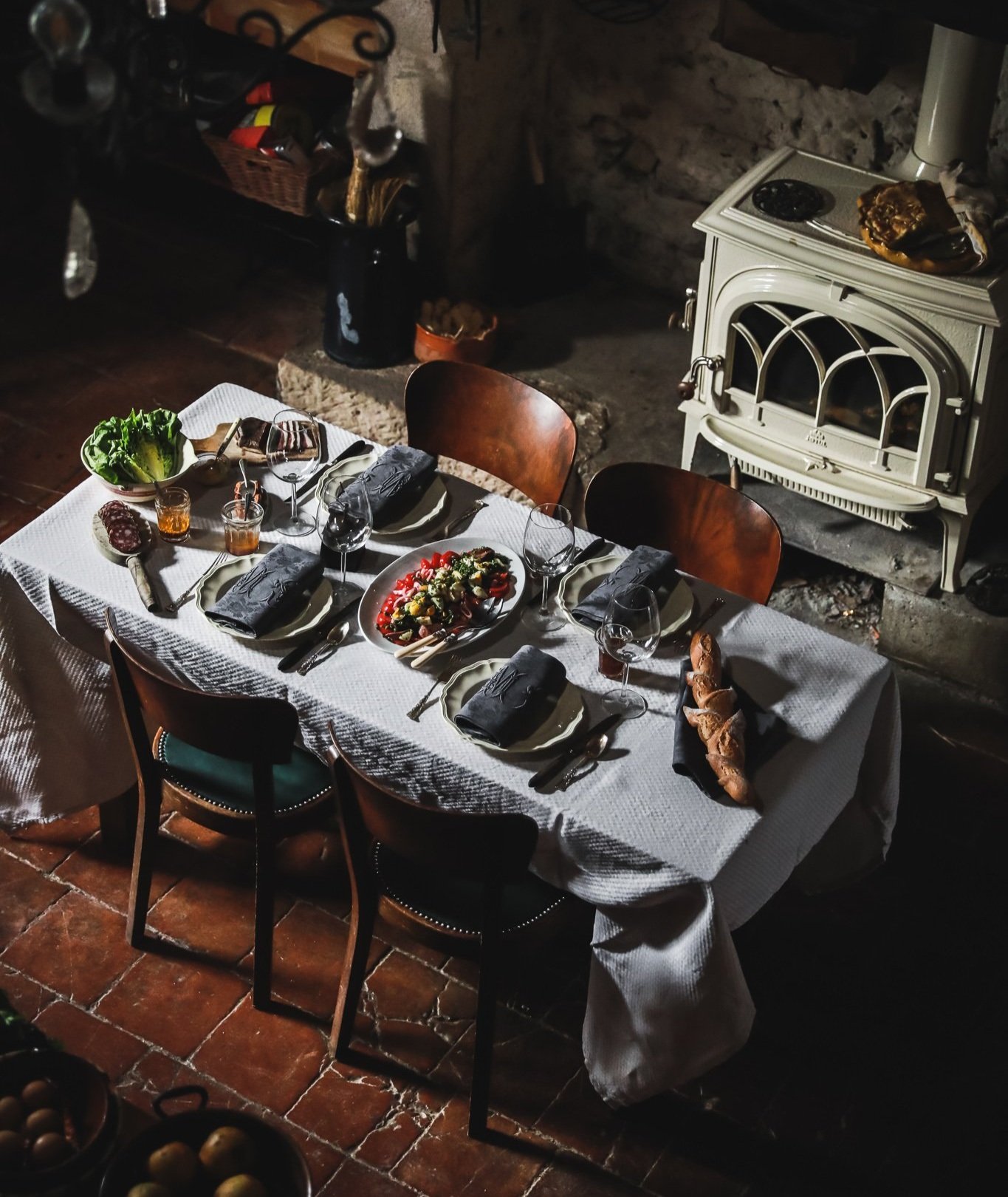 A rustic dining table set for a meal, with plates, glasses, cutlery, a salad, bread, and napkins, in a cozy, dimly-lit room with a brick fireplace and vintage oven.