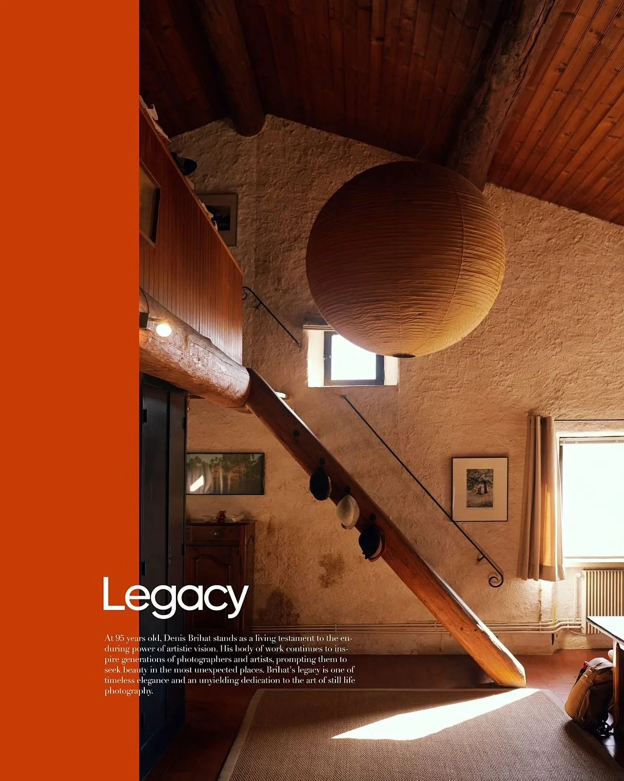 Interior of a rustic room with wooden ceiling and beams, a large paper lantern hanging from the ceiling, small windows allowing natural light, and a staircase with hats hanging on the handrail. Sunlight casts shadows on the wall and floor.