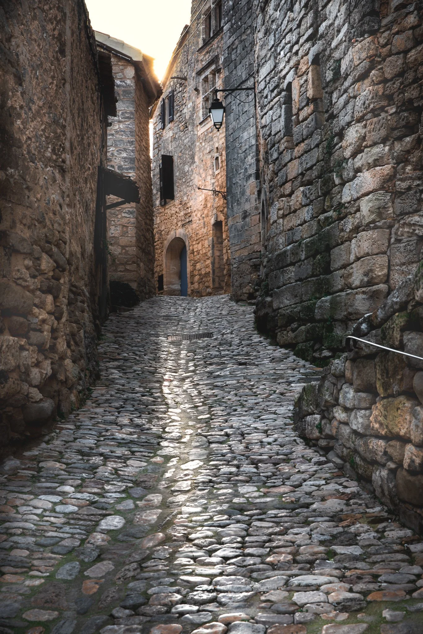 A narrow cobblestone alleyway between tall, historic stone buildings with a lantern hanging from a wall.