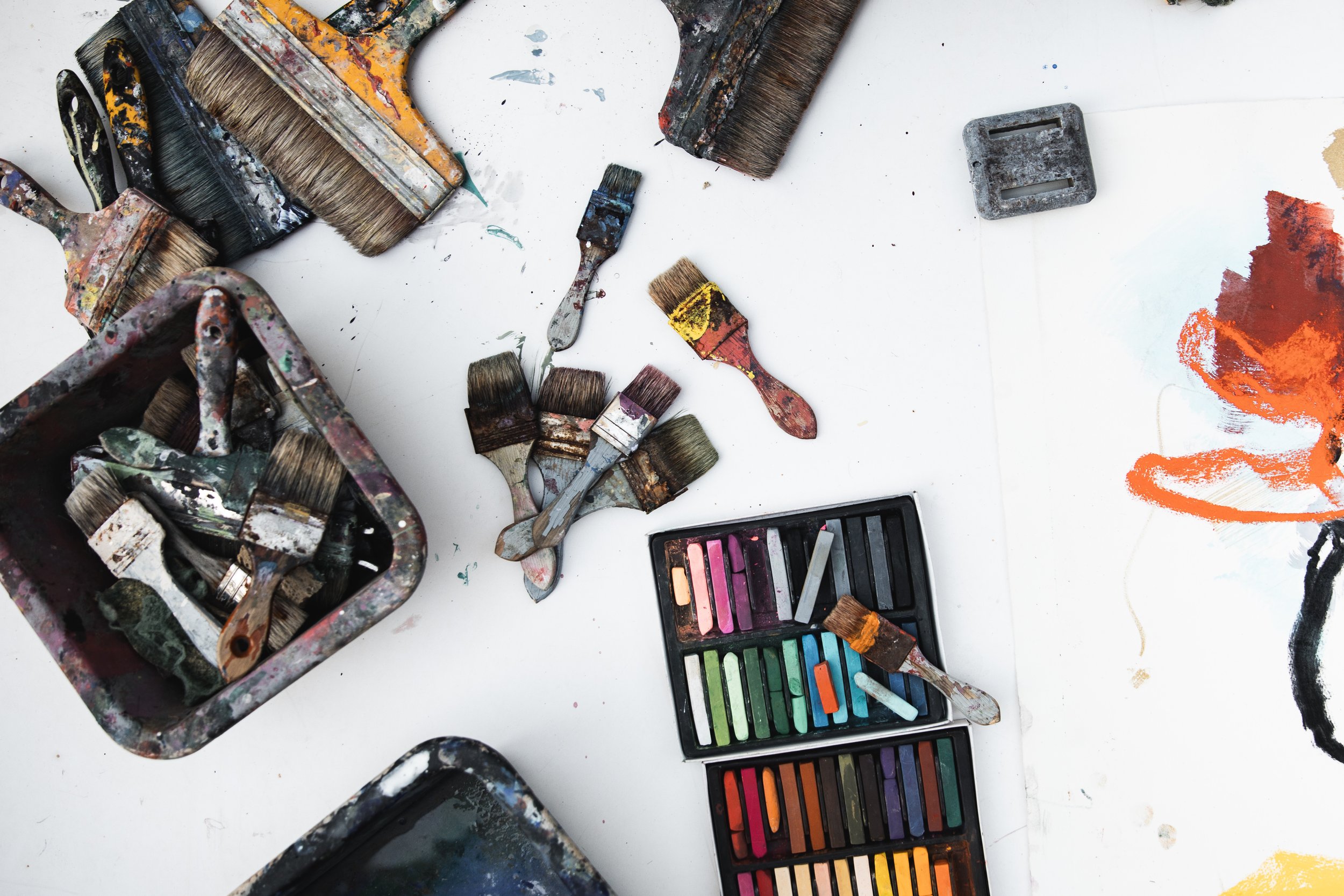 Assorted paintbrushes, paint bottles, and pastel chalks scattered on a white surface.