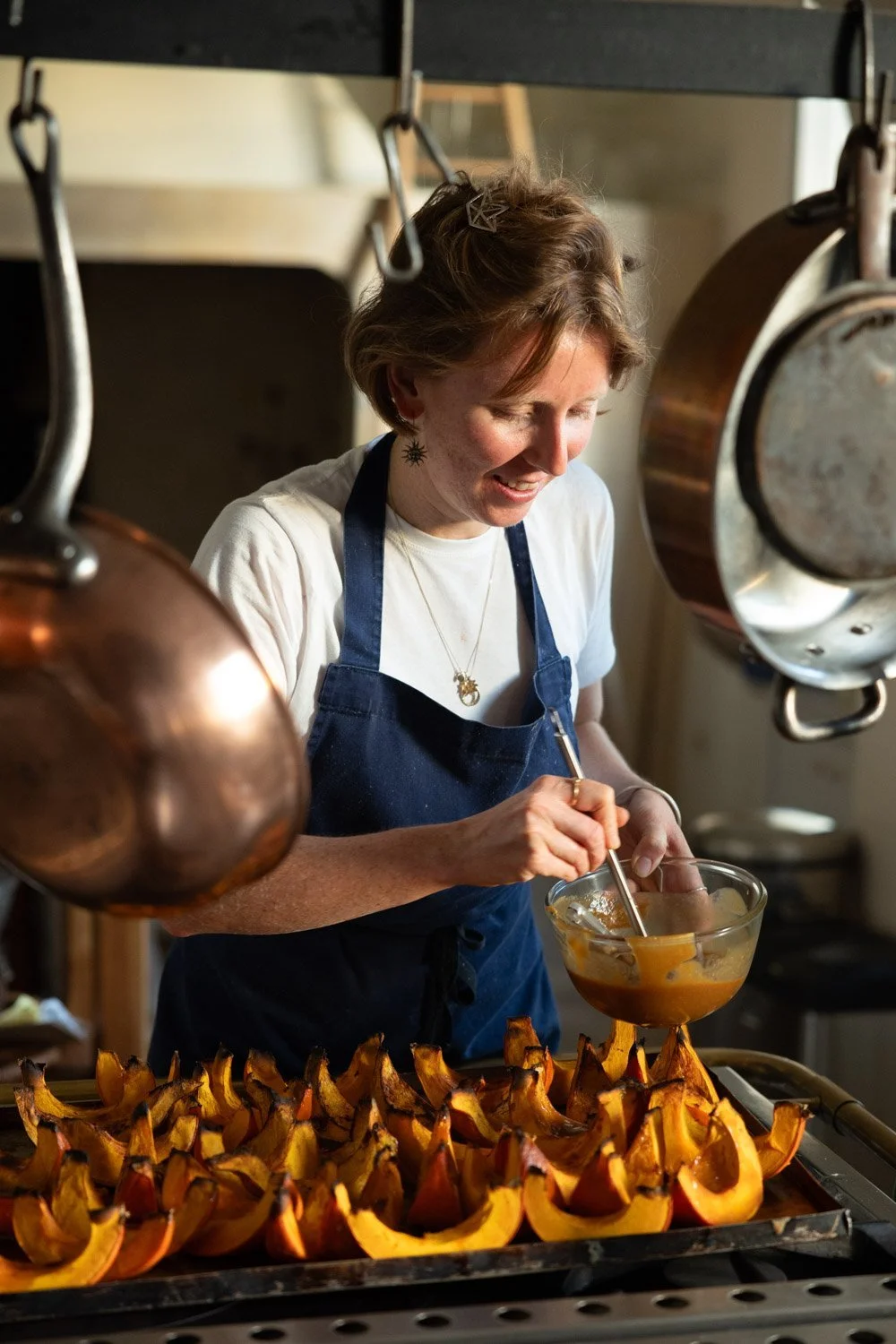 Woman in a navy apron mixing pumpkin in a glass bowl in a kitchen, with roasted pumpkin halves on a tray in front of her.