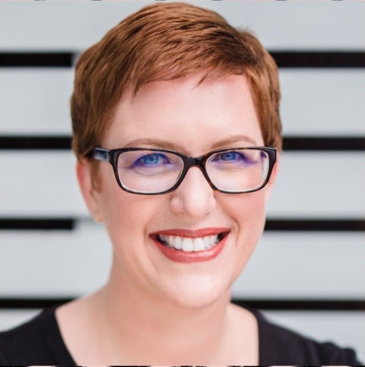 Close-up of a smiling woman with short red hair, black glasses, and a black top, standing in front of a background with horizontal black and white stripes.