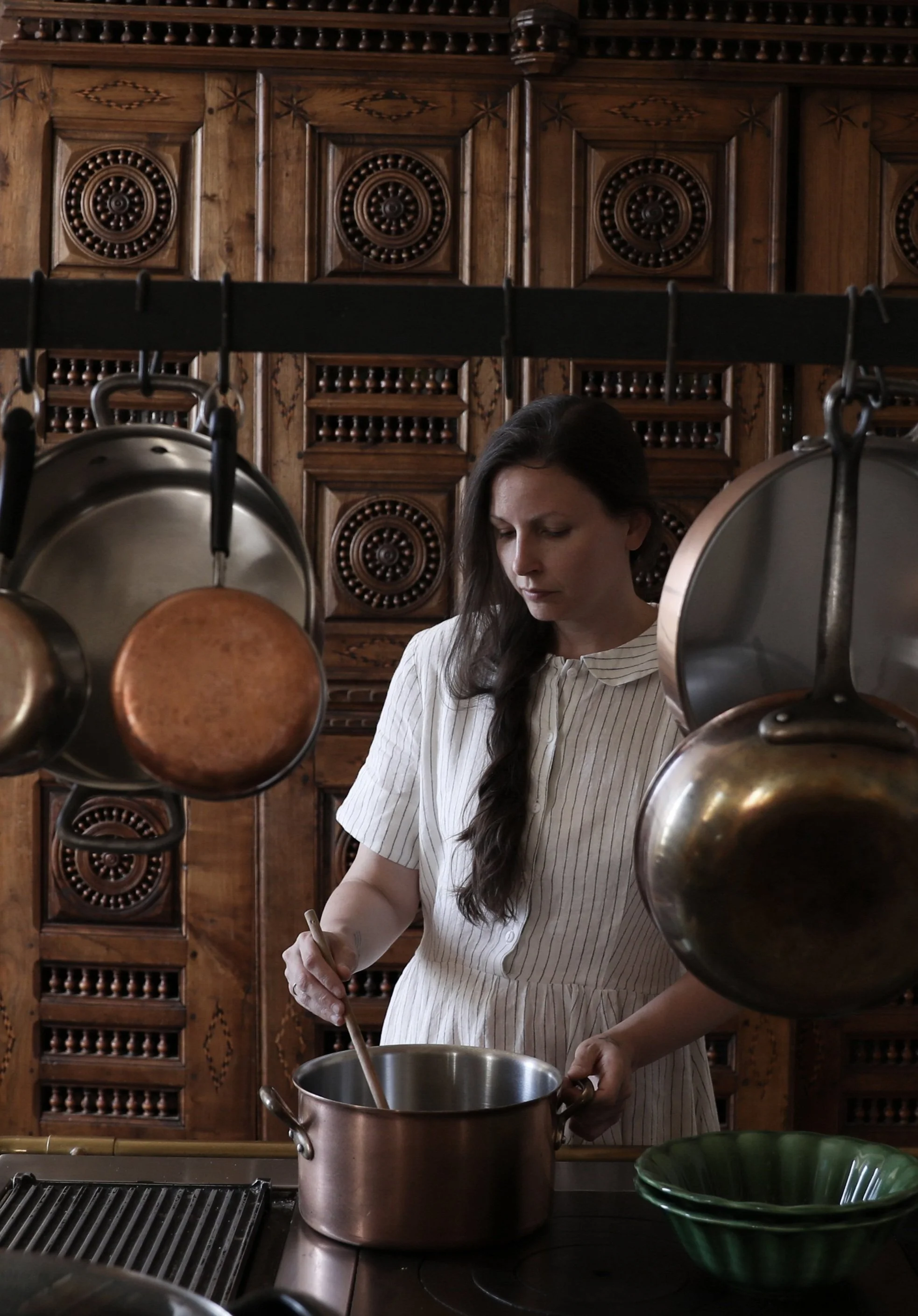 A woman with long dark hair, wearing a white and beige striped dress, cooking in a kitchen with ornate wooden cabinets, hanging pots, and a black stove, stirring a pot.