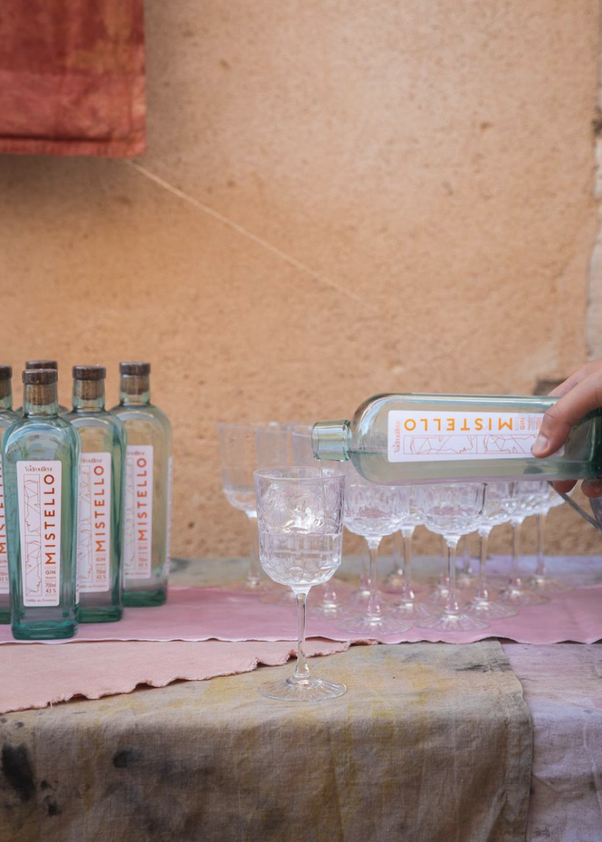 A person is pouring Mistello gin into a glass of ice on a table with a pink cloth, with several other glasses and bottles of Mistello gin in the background.