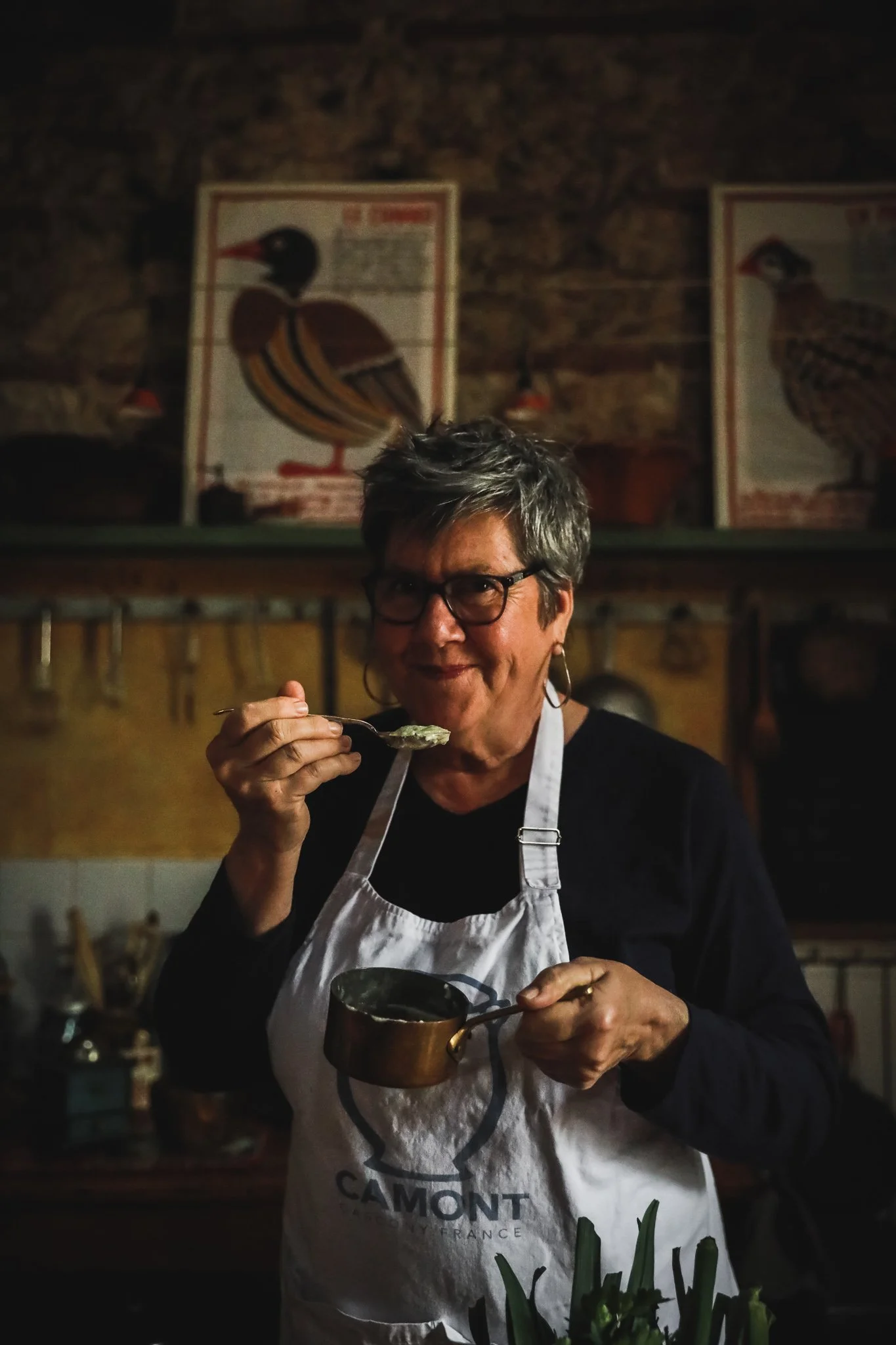 A woman with short gray hair, glasses, and hoop earrings, wearing a white apron with "CAMONT" printed on it, is smiling while holding a spoon with food, standing in a kitchen with arts and craft items on the walls.
