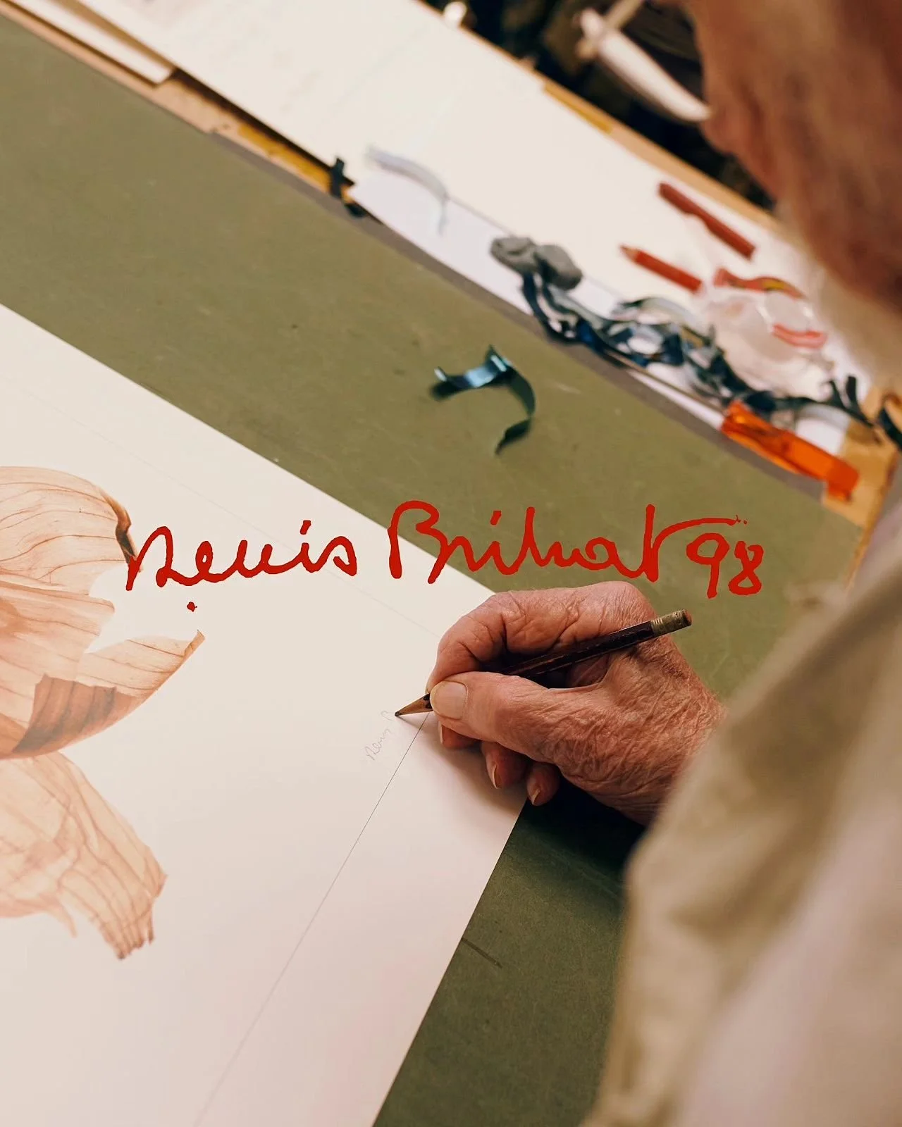 An elderly person signing a poster with a black pen. The poster has a leaf illustration and a signature that reads 'Lewis Bihart 98' in red. The workspace has a green cutting mat, scissors, and other tools in the background.