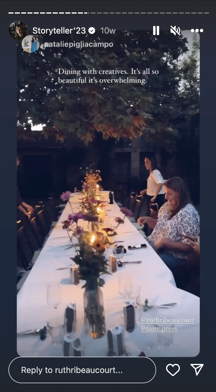 An outdoor dinner party with a long table decorated with flowers and candles, under a tree with autumn leaves, with people standing and sitting around the table.