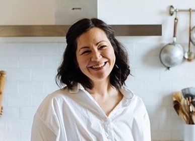 Woman with dark hair smiling in a kitchen with white tiles and wooden accents.