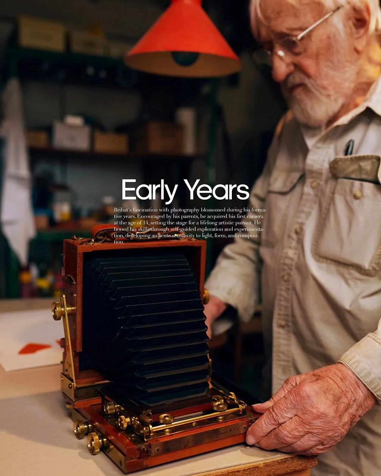 An older man with gray hair and glasses, wearing a beige shirt, working with a large, vintage camera with a bellows in a workshop or studio.