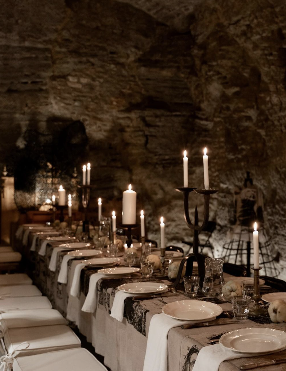 Long dining table set with white plates, forks, glasses, and white cloth napkins, decorated with tall white candles in candleholders, inside a cave with a rocky wall background.