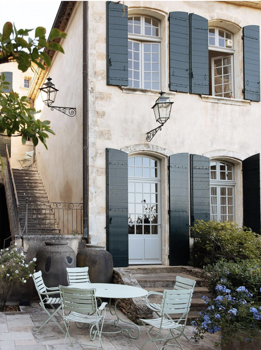 A quaint outdoor patio with white metal table and chairs, large clay pots, lush green bushes, and a building with beige stone walls, dark green shutters, and three windows.