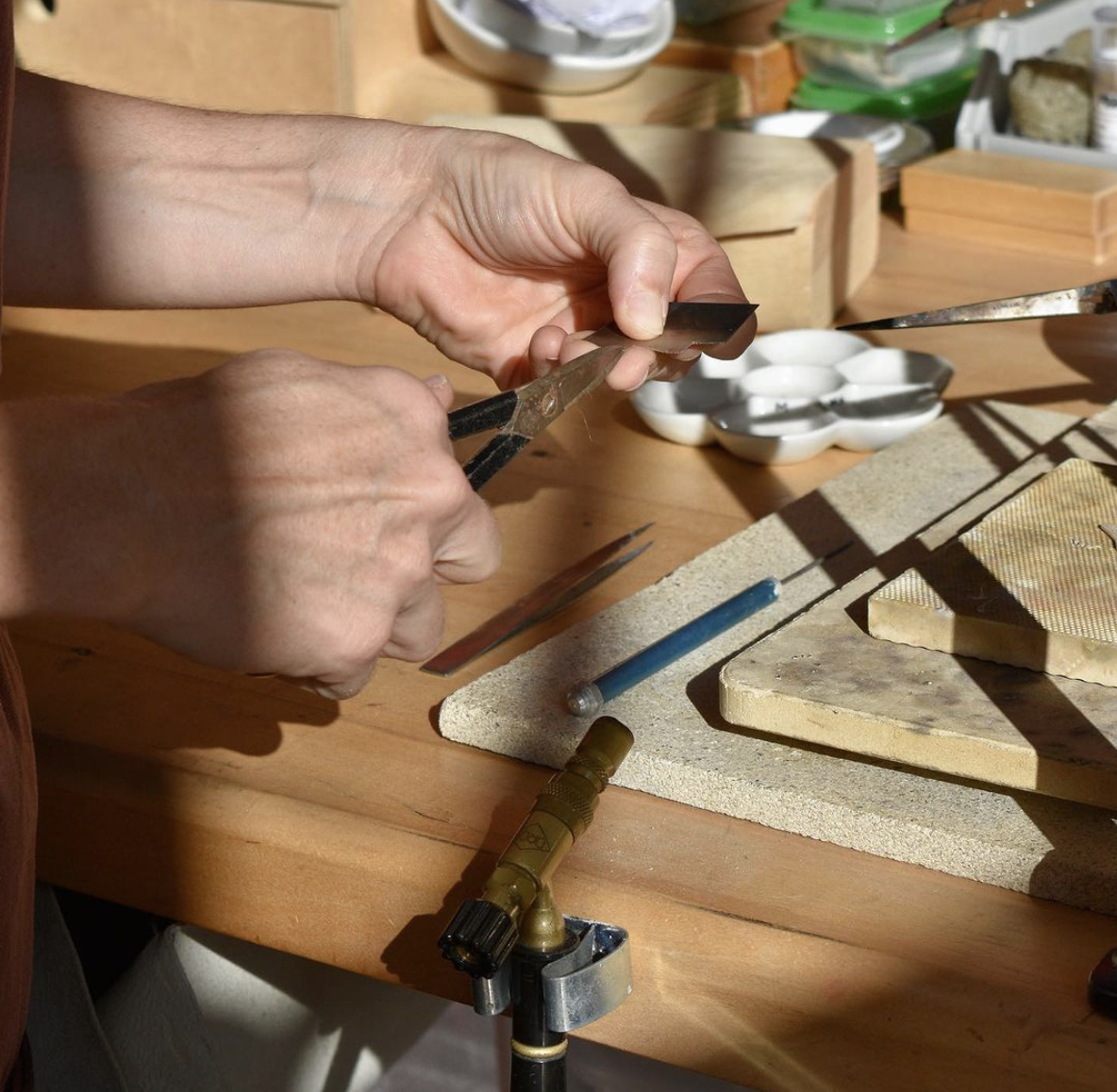 Hands working with jewelers tools on a piece of jewelry in a workshop with various small tools and containers on a wooden workbench.