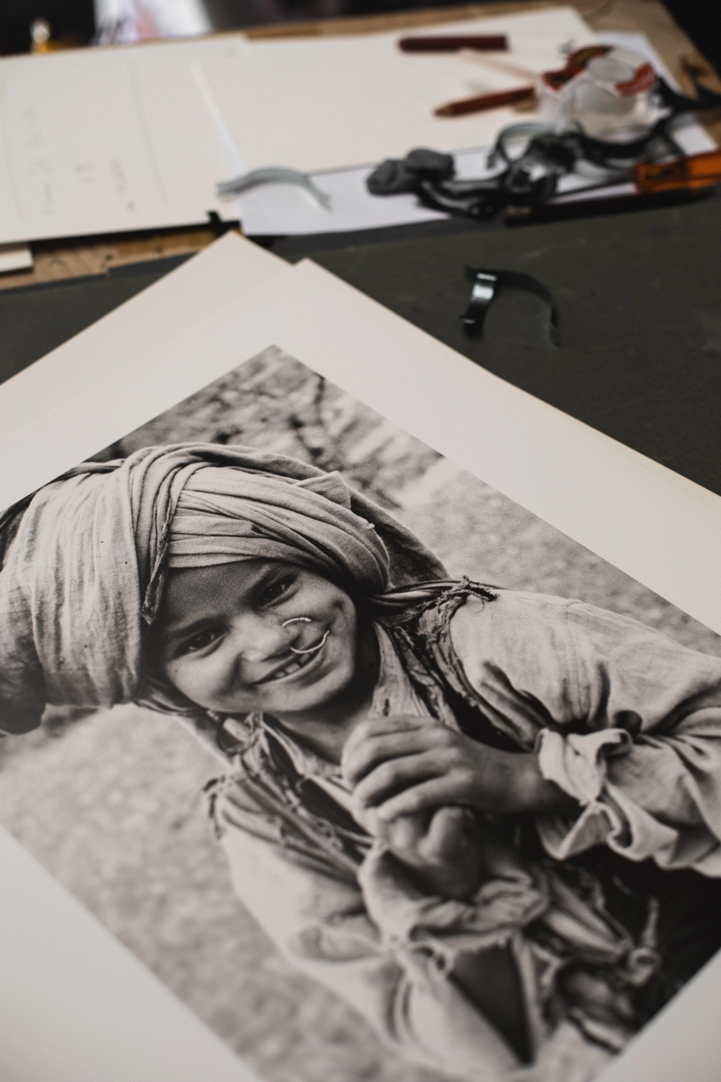 Black and white portrait of a smiling child wearing a headscarf and jewelry, outdoors with trees in the background.
