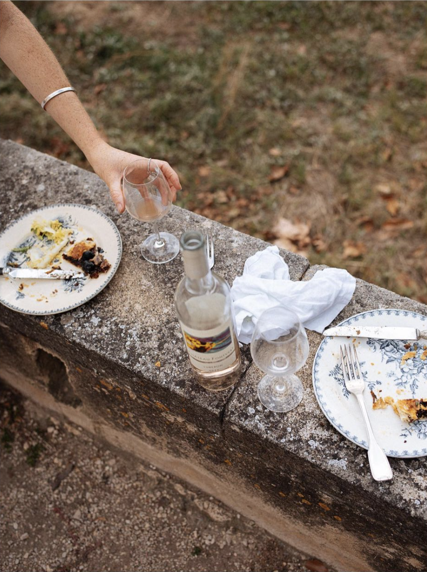 Empty dinner plates with partially eaten cake, two empty wine glasses, a bottle of rosé wine, and a crumpled white cloth on a stone ledge outdoors.