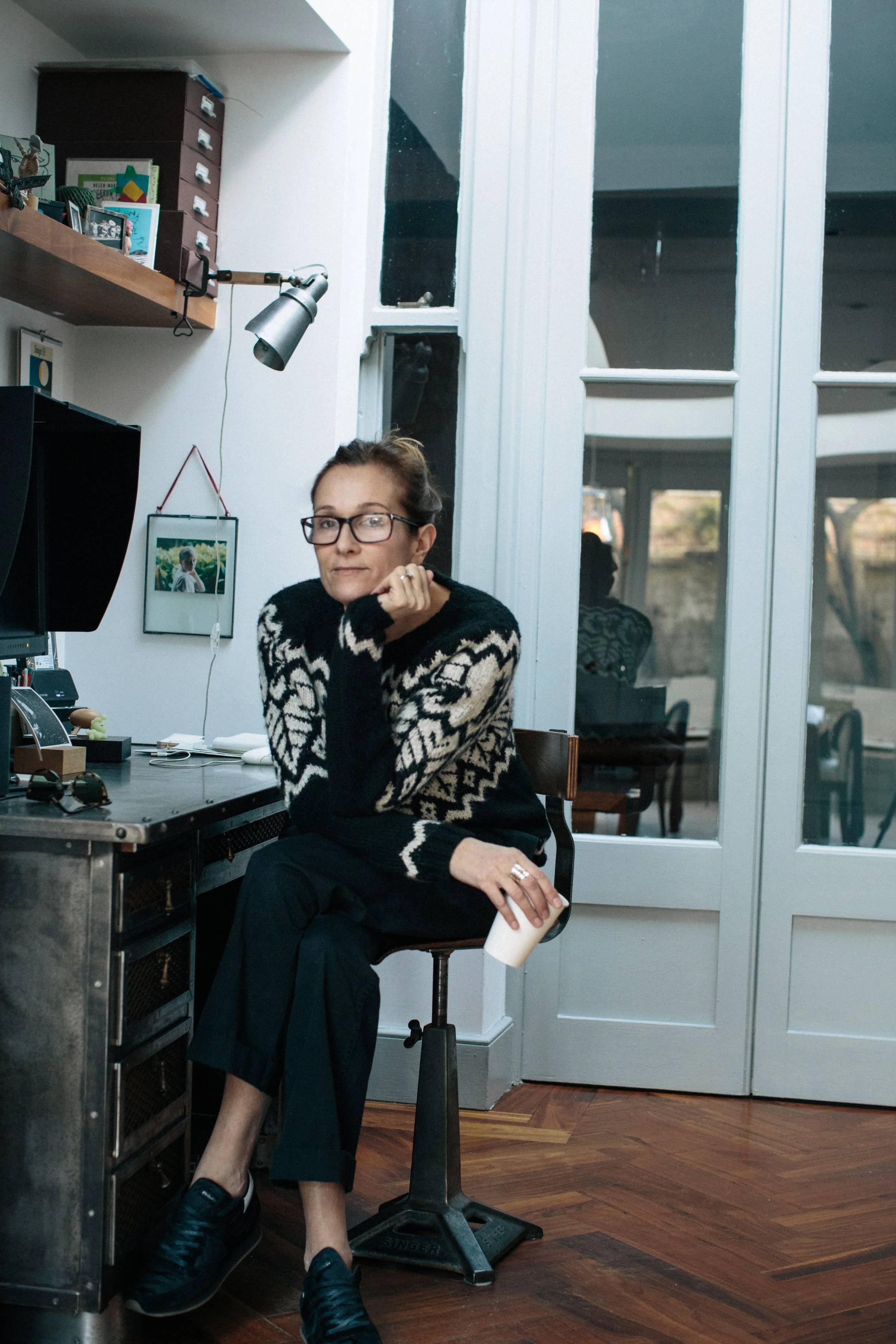 A woman with glasses sitting at a desk in a home office, resting her chin on her hand, holding a cup in her lap, with a workspace, computer, and photos on the wall behind her.