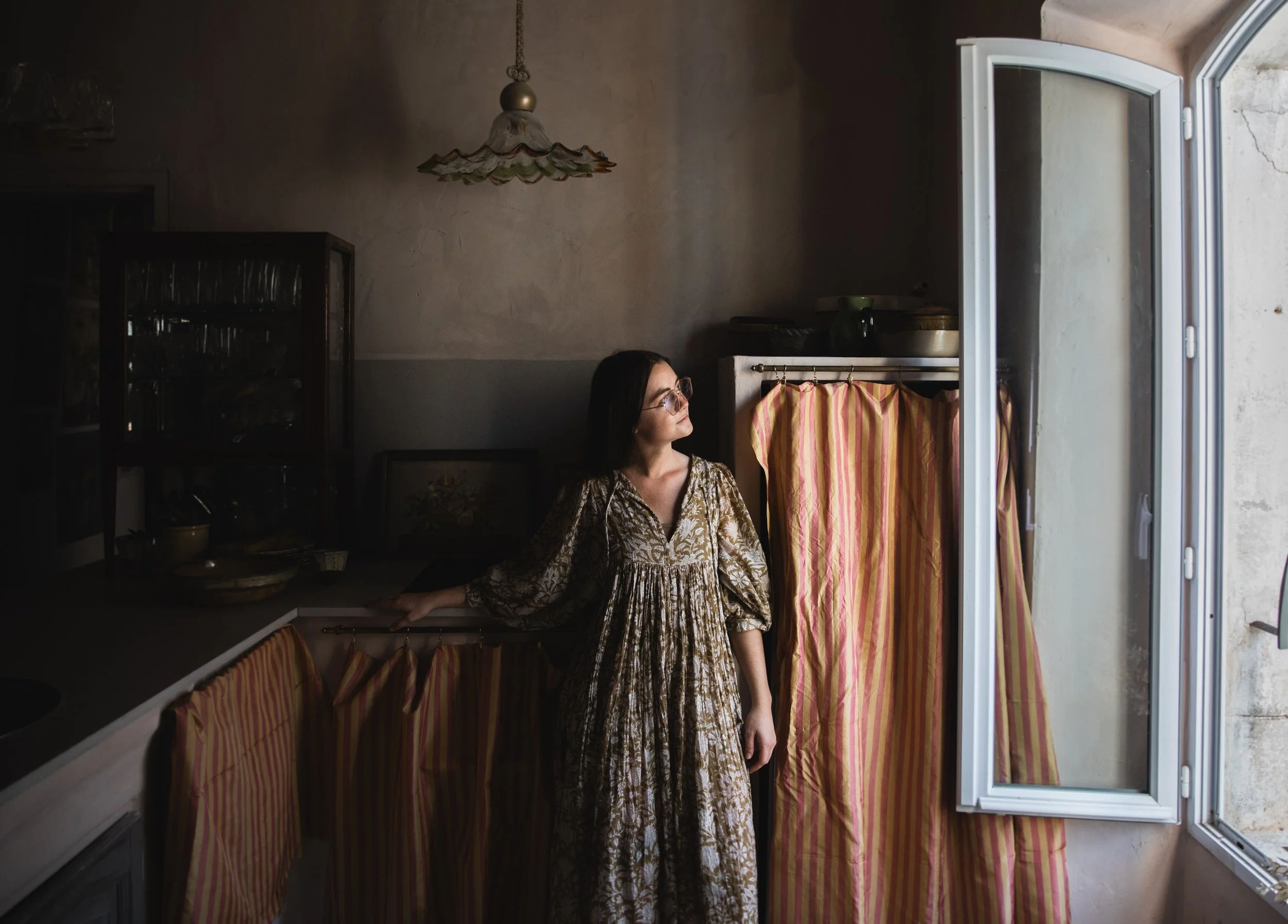 Woman in vintage dress standing in a dimly lit kitchen near an open window with red and yellow striped curtains, wearing glasses and looking to the side.