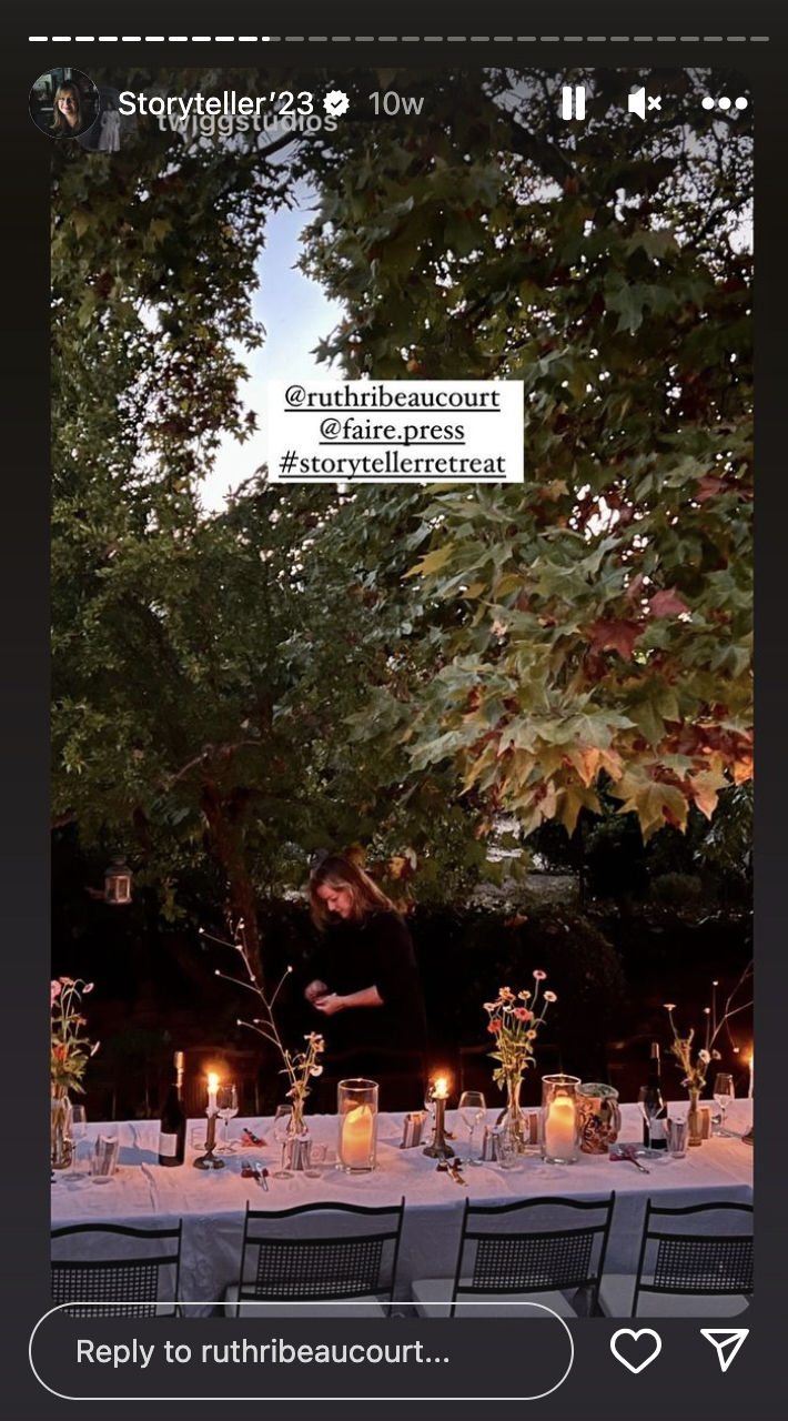 An outdoor dinner setup under a tree with a long table decorated with candles and flowers. There is a woman standing behind the table, possibly preparing or setting the table, with a backdrop of trees and foliage.