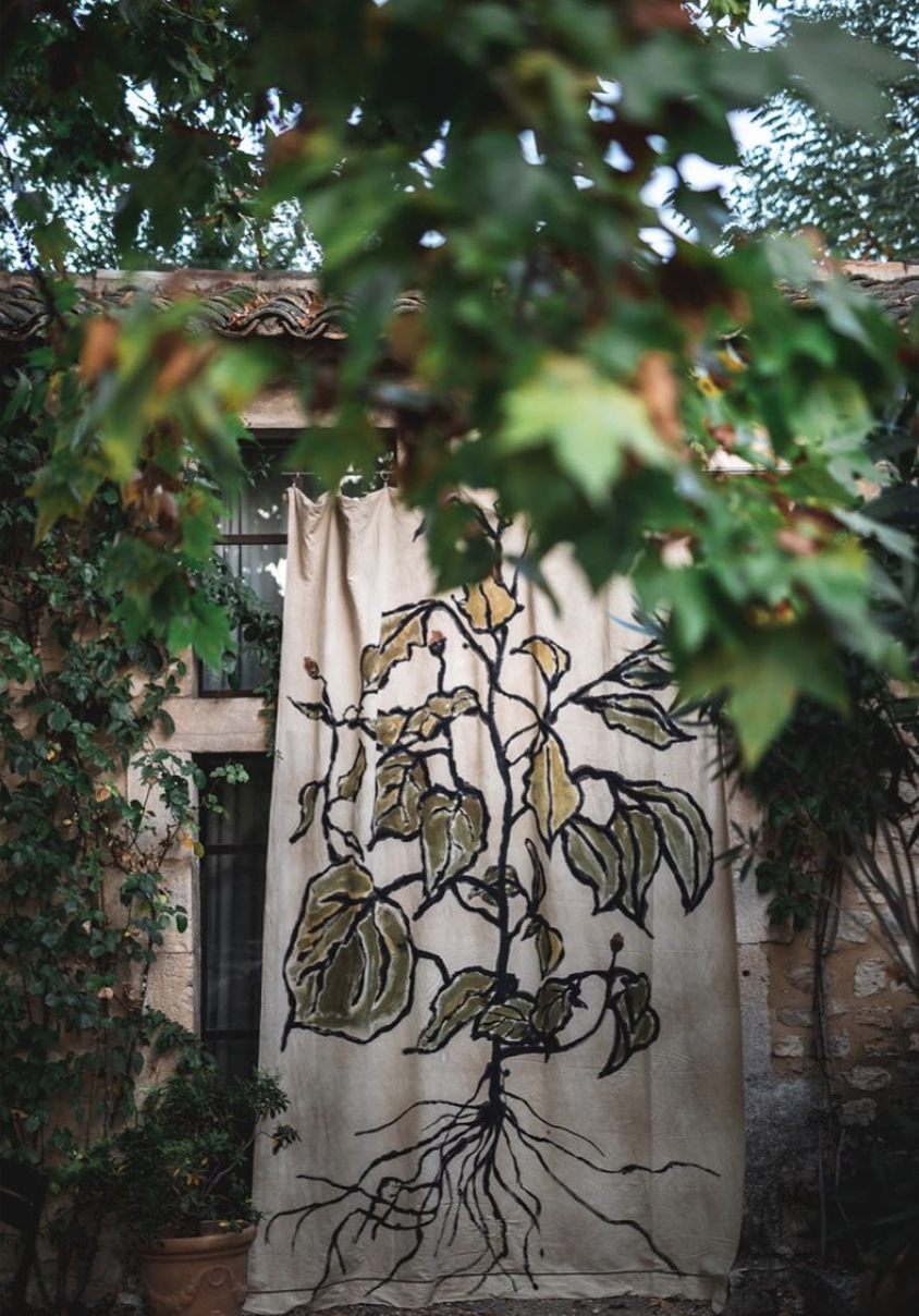 A decorative fabric or curtain with an illustration of a plant's roots and leaves, hanging outdoors against a stone wall, surrounded by lush green foliage.