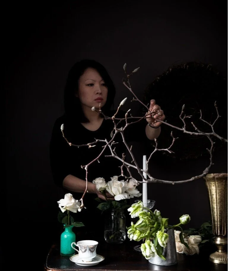 A woman arranging flowers and branches on a dark background with various vases and a teacup on a table.