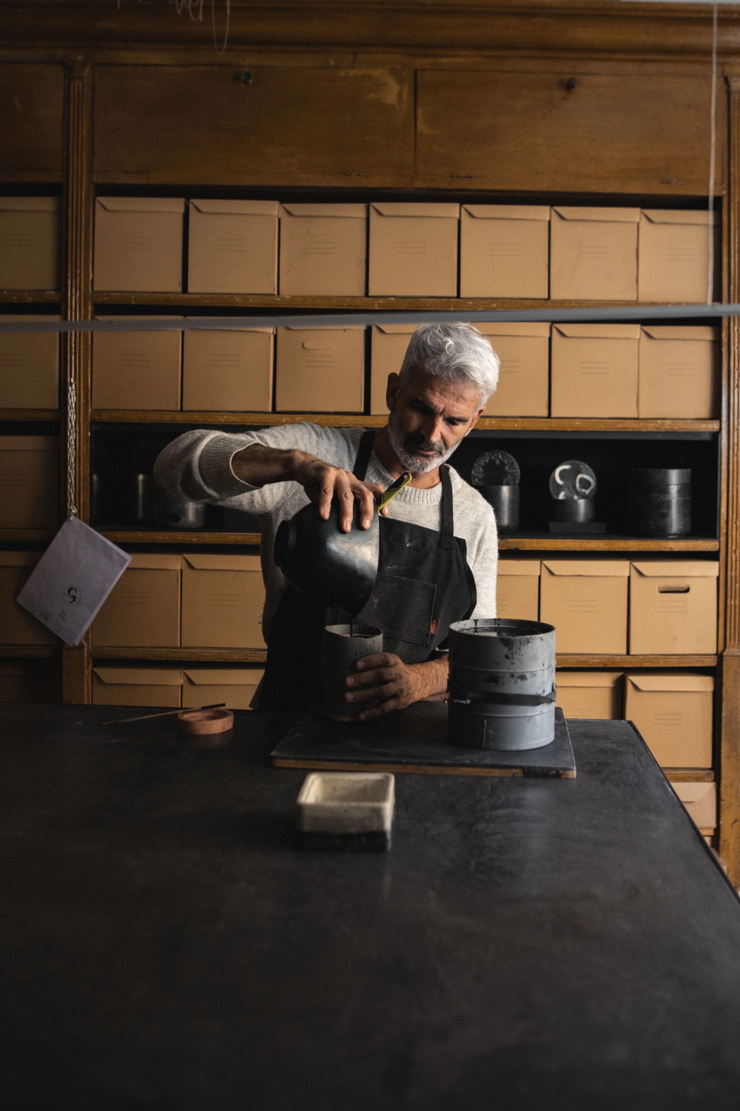An elderly man with gray hair and a beard pouring dark liquid into a cup inside a rustic kitchen with wooden shelves and cardboard boxes.