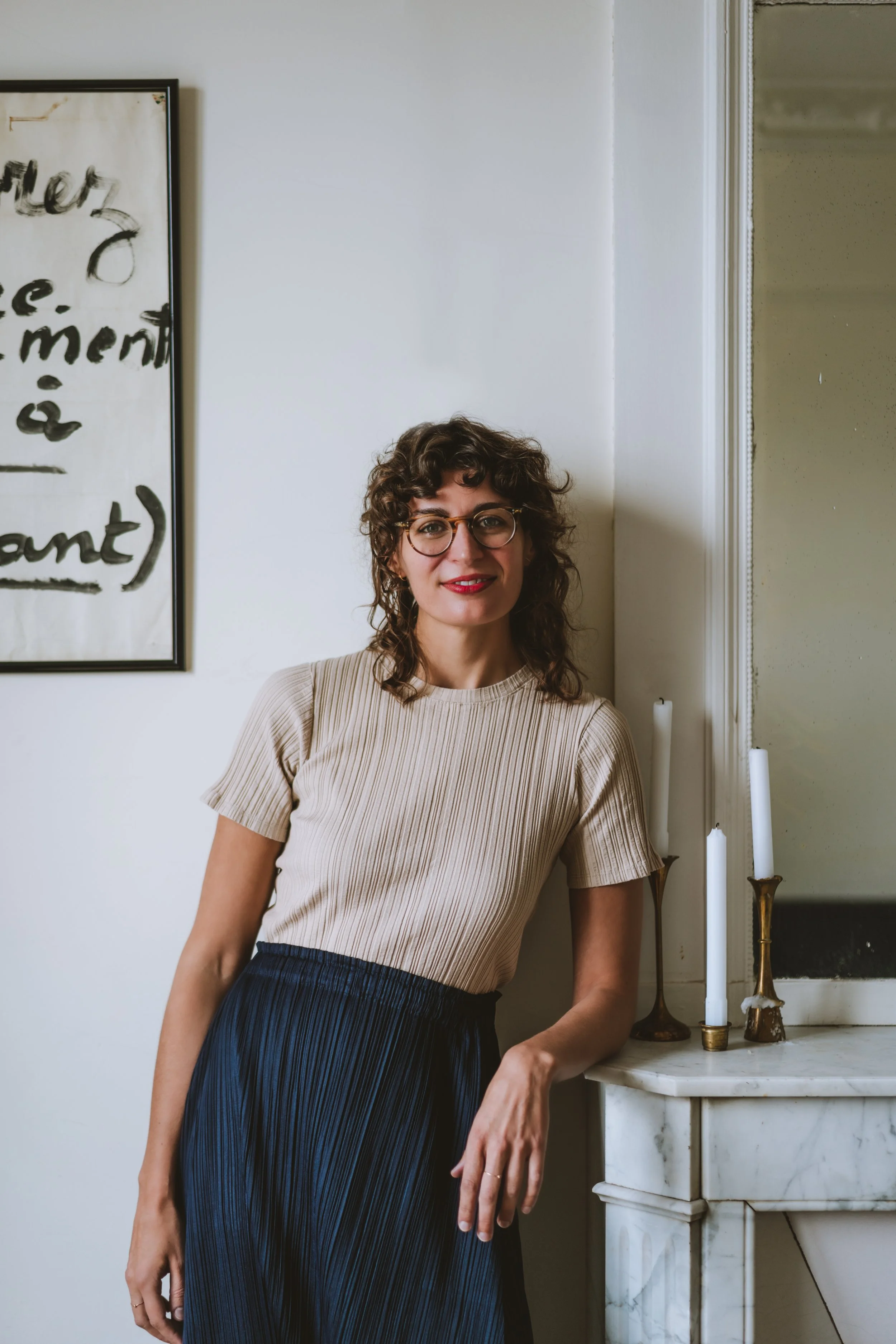 A woman with curly brown hair, glasses, and red lipstick, wearing a beige ribbed top and a dark pleated skirt, standing next to a marble fireplace with three white candles in brass holders. She is smiling and leaning slightly on her right arm.