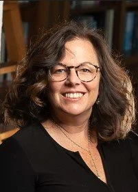 A woman with curly brown hair, glasses, and a black top, smiling in an indoor setting.