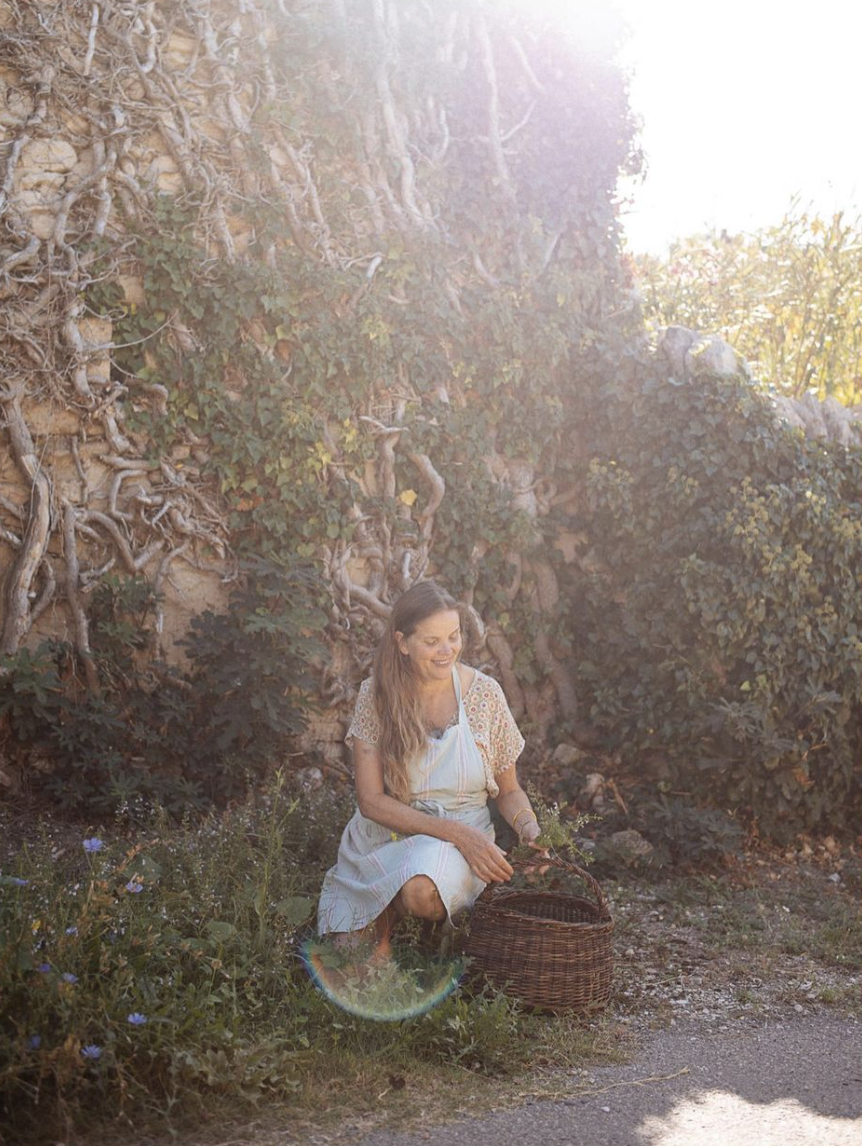 A woman in a light-colored dress kneels on the ground, smiling while gathering flowers with a wicker basket beside her, in front of a vine-covered stone wall and bushes, with sunlight creating lens flare.