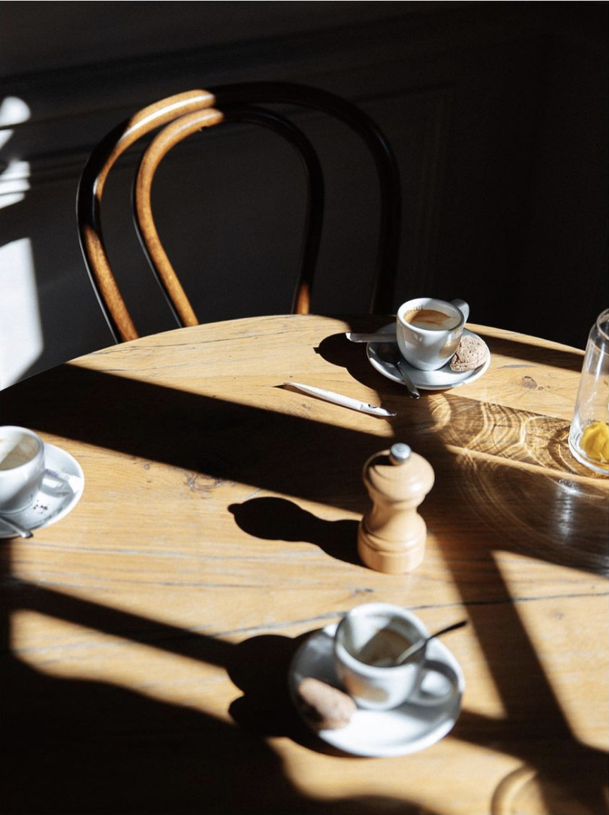 A wooden table with three white coffee cups on saucers, a small spoon in each cup, and two cookies on the saucers. A black pepper grinder is in the center, casting a shadow. Natural sunlight creates dramatic shadows across the table and a dark backgr