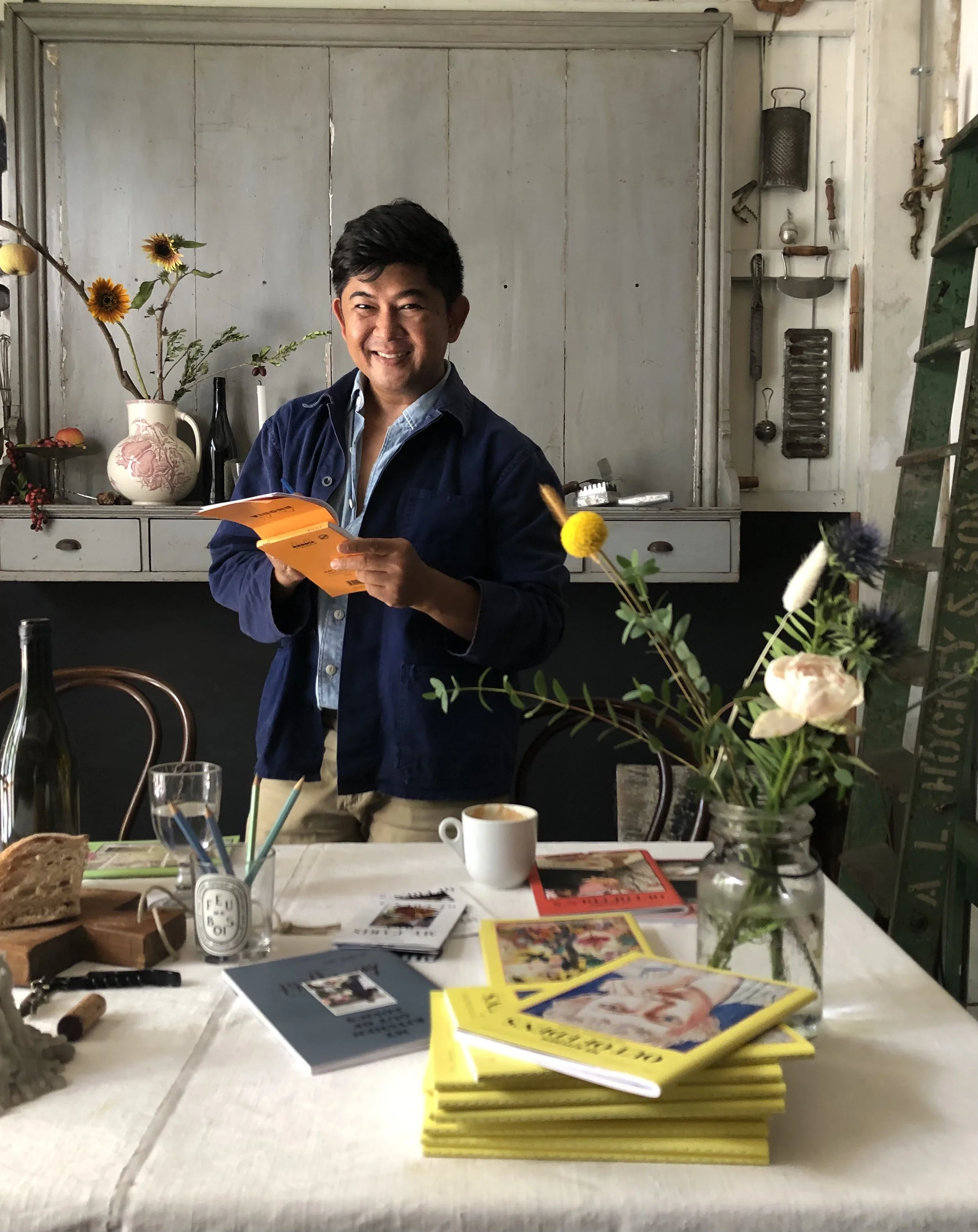 A smiling man reading a booklet in a cozy, rustic room with a table set with books, a mug, and flowers in a jar.
