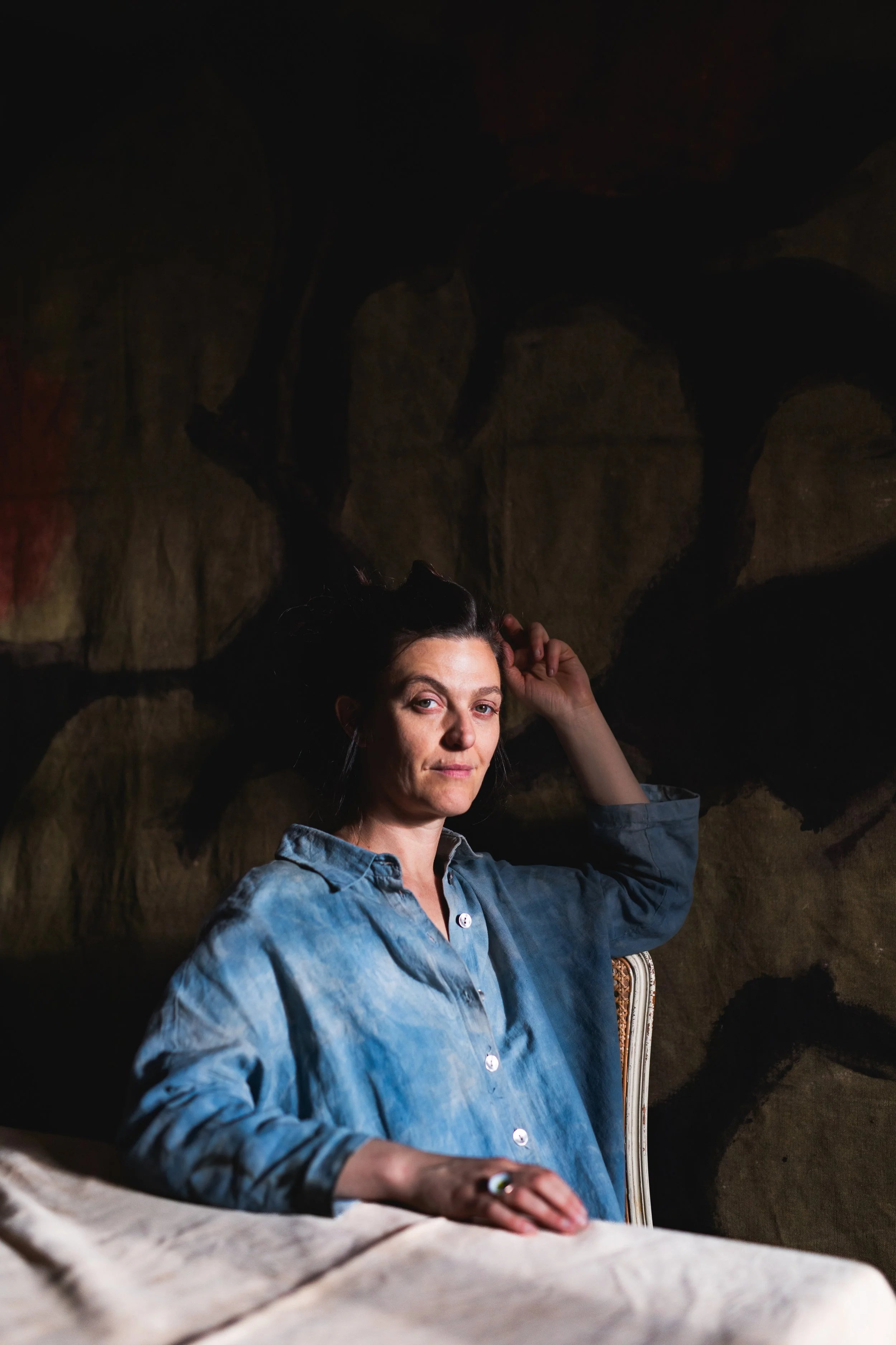 A woman with dark hair wearing a blue button-up shirt sitting in front of a dark, textured background with abstract shapes.
