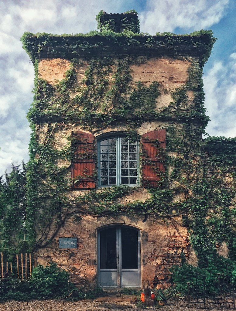 An old brick and stone building with green ivy covering parts of the facade and red wooden shutters on the windows. The structure has a rounded door at the bottom and a larger arched window above. The building appears to be in a garden or yard with plants and a blue sky with clouds in the background.