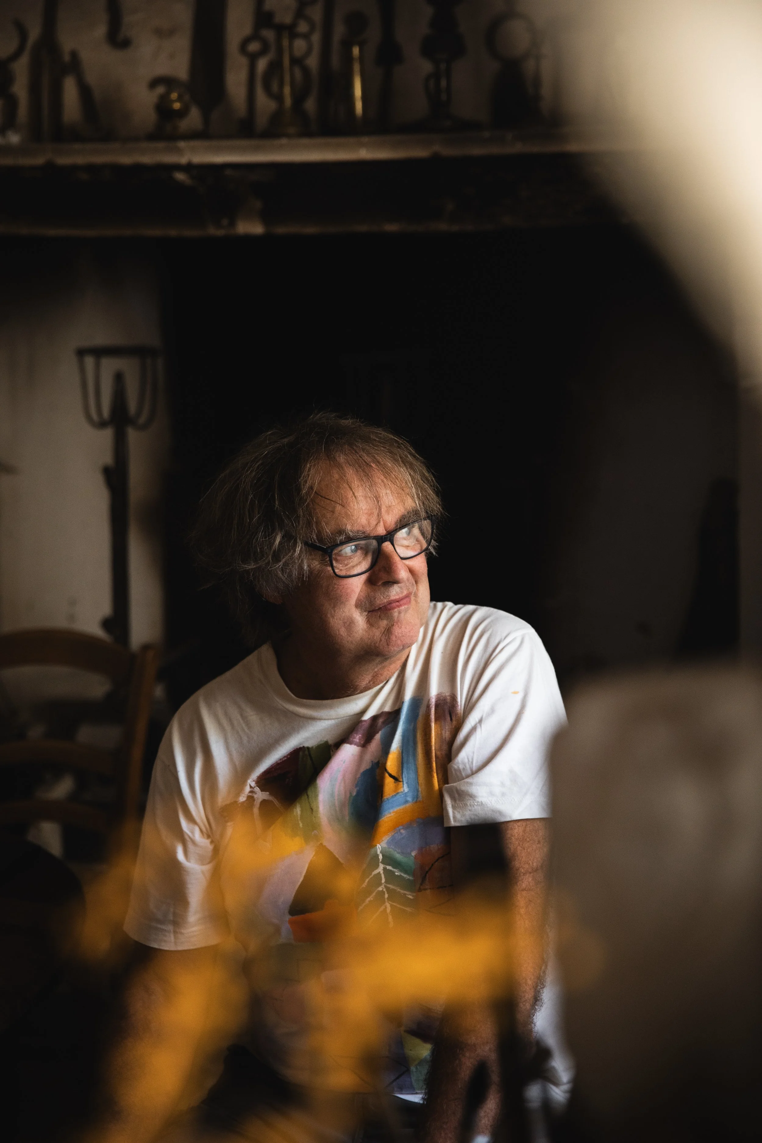 A man with messy hair and glasses sitting indoors, wearing a colorful graphic T-shirt, looking to his right. In the background, there are chairs, a fireplace or dark opening, and shelves with various objects or decorations.