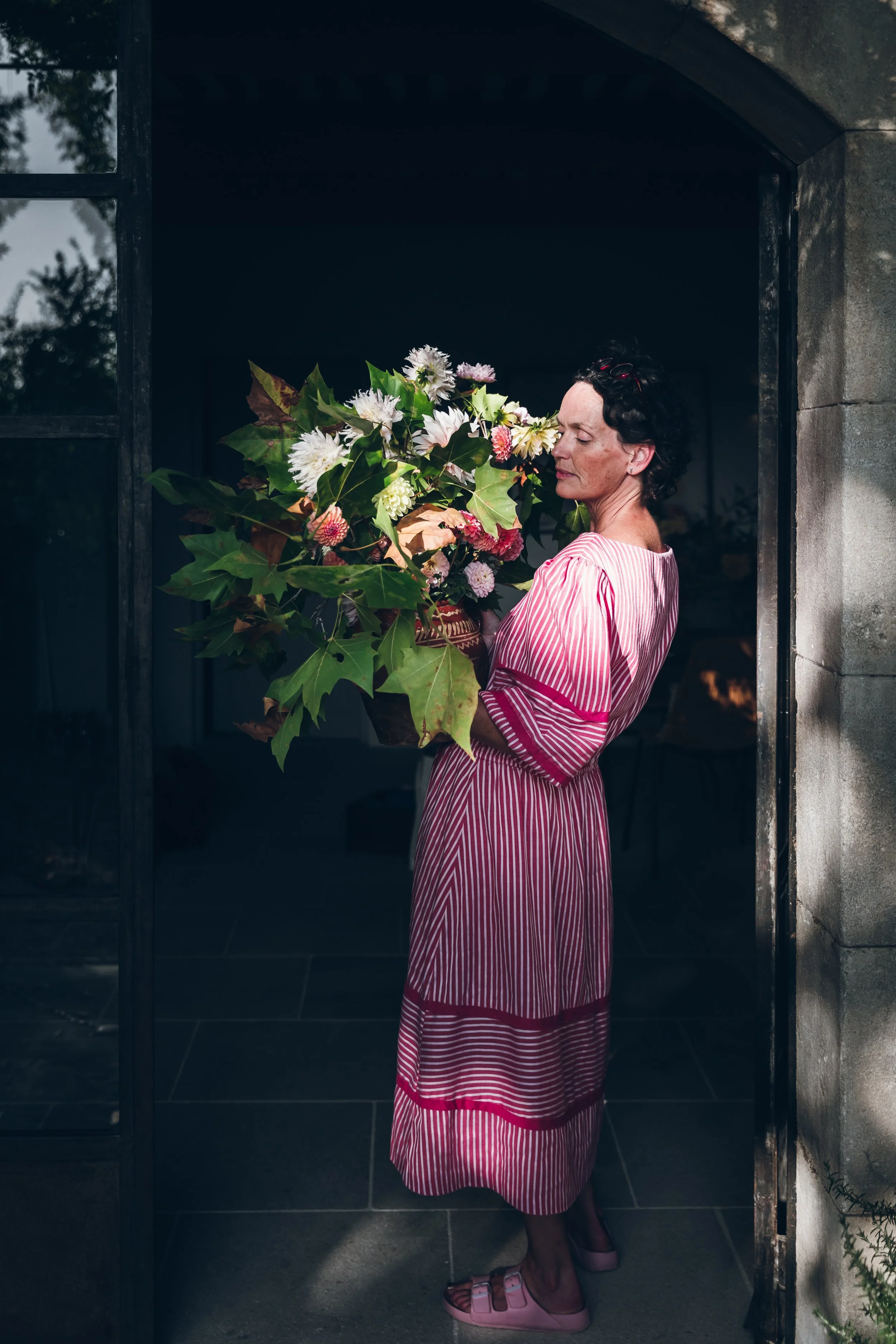 A woman in a pink and white striped dress holding a large bouquet of flowers and greenery near a stone archway.