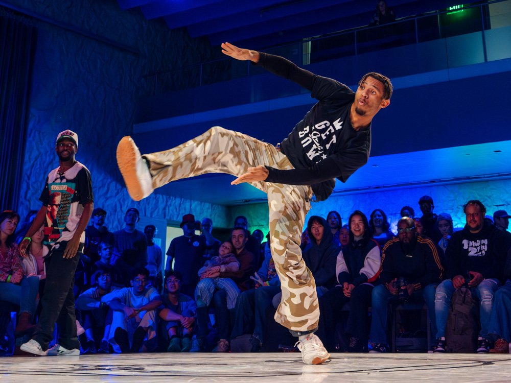 A dancer moves upright with rhythmic footwork and expressive arm movement in a hip-hop freestyle cypher, surrounded by seated spectators.