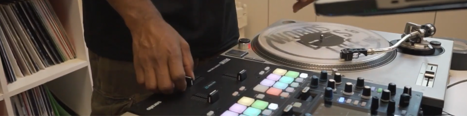 Person's hand operating buttons on a music production controller next to a turntable with vinyl records in background.