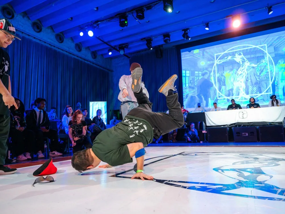 A dancer balances on one arm in a breaking freeze inside a cypher, with an audience gathered closely around the floor.