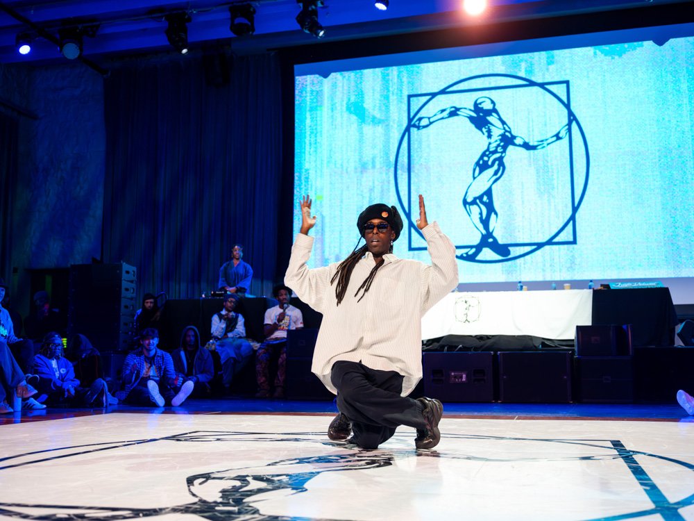 A dancer performs house footwork with an upright posture while spectators sit and stand around the dance floor.