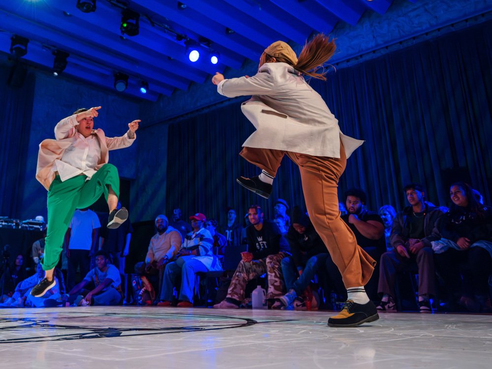 Two dancers face each other in a house dance exchange, moving low to the floor as the audience watches from a circle.