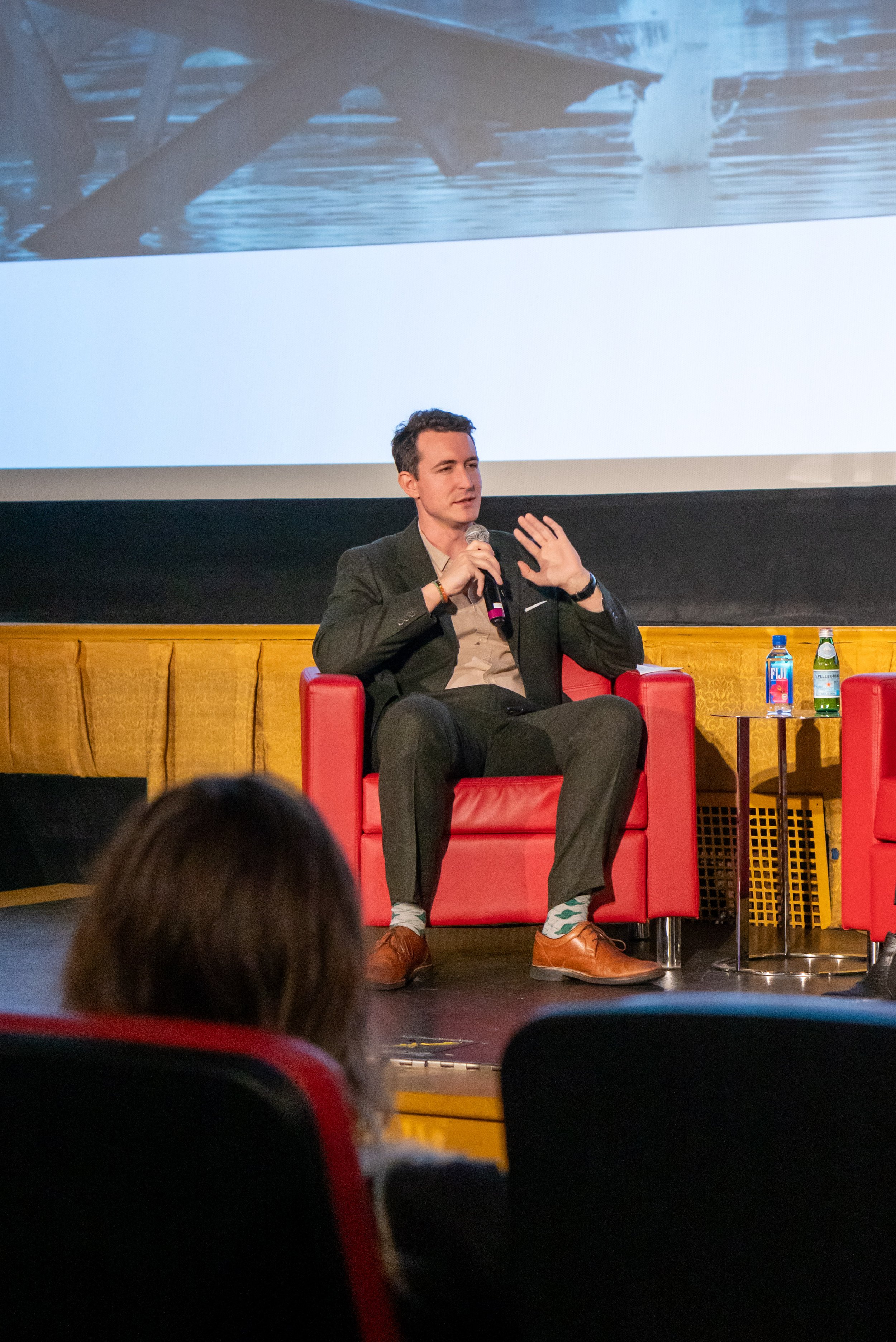A man in a dark suit sits in a red armchair on stage, speaking into a microphone. Behind him is a large screen displaying an image of a wooden dock over water. To his right, there is a small table with bottled water.