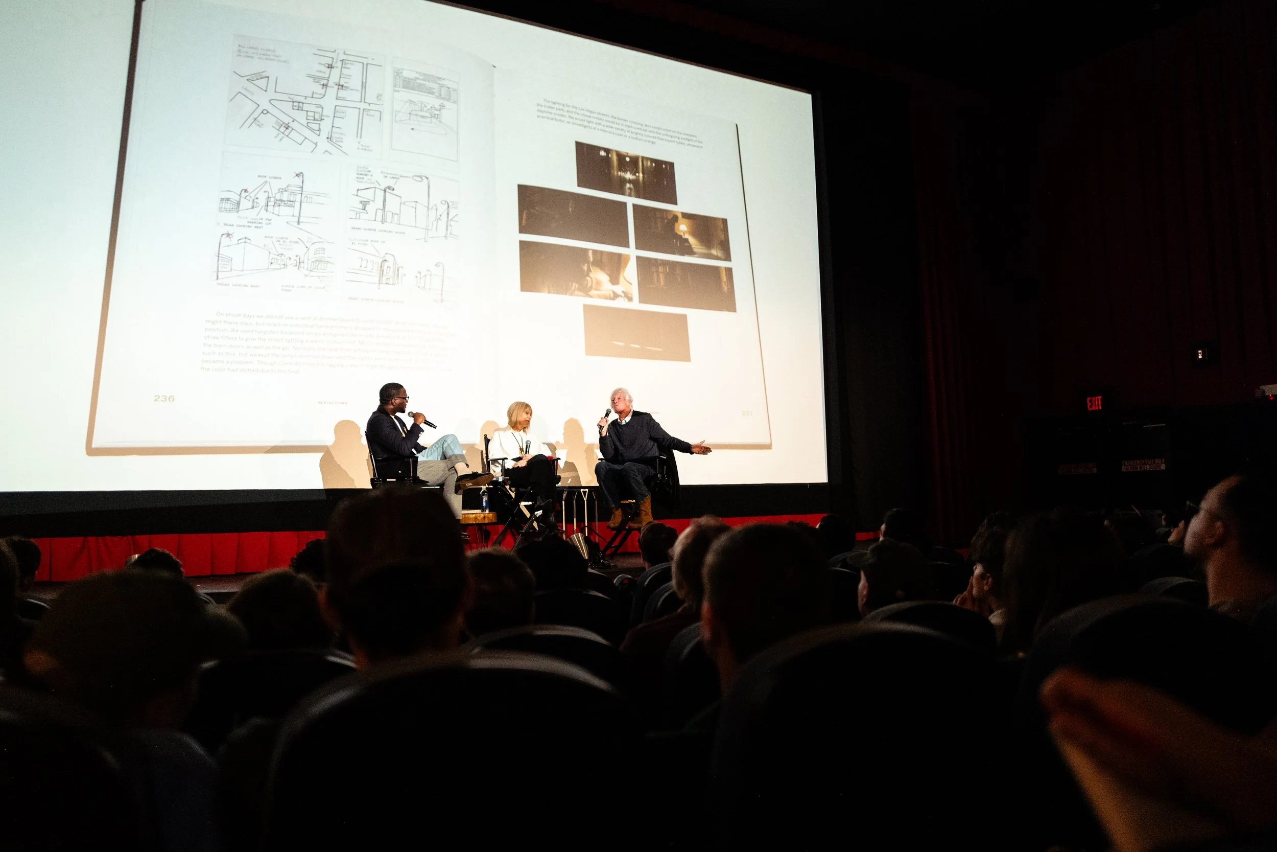 A panel discussion on stage in a theater with a large screen behind, showing presentations and images. Three people are sitting on chairs, speaking into microphones, engaging with the audience.