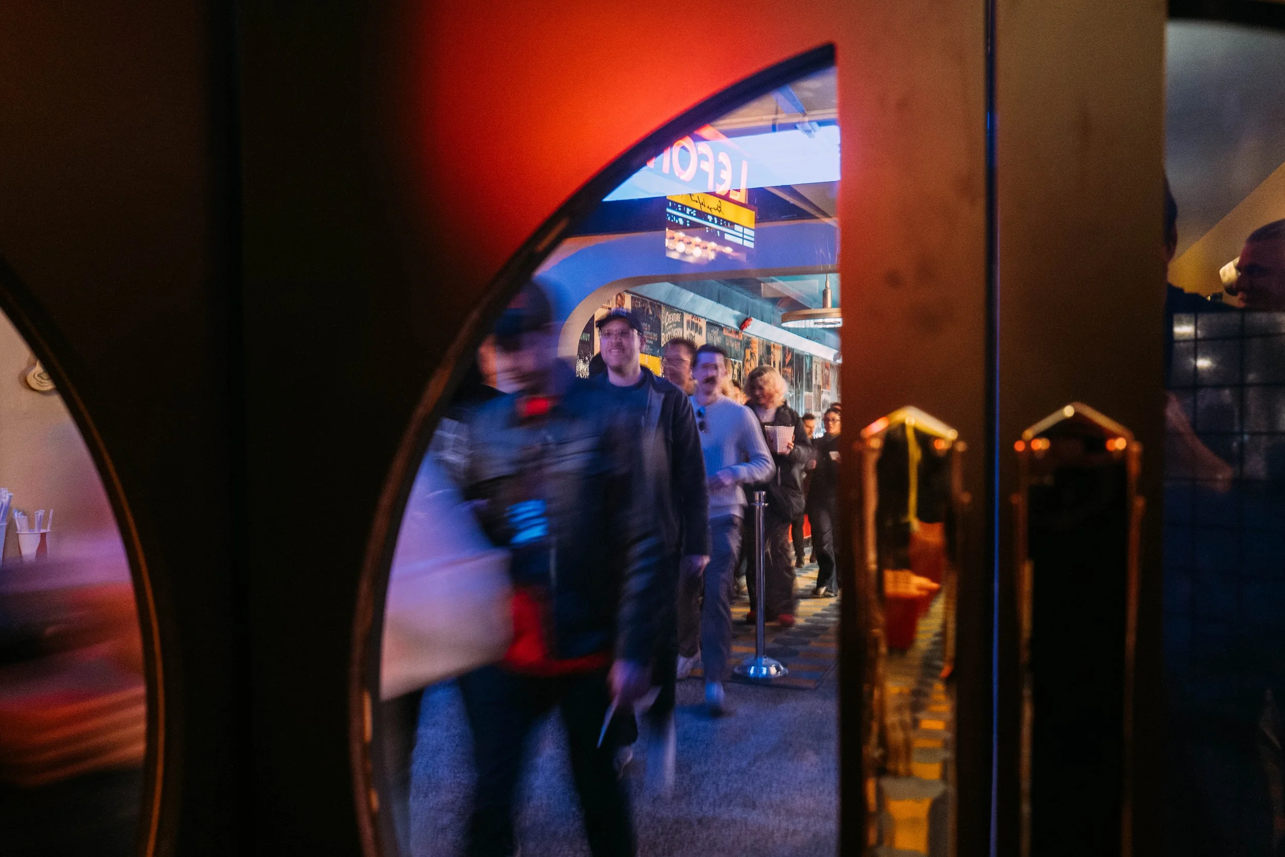 People standing in line at a venue, seen through an arcade game mirror, with some smiling and others looking forward, illuminated by colorful lighting.