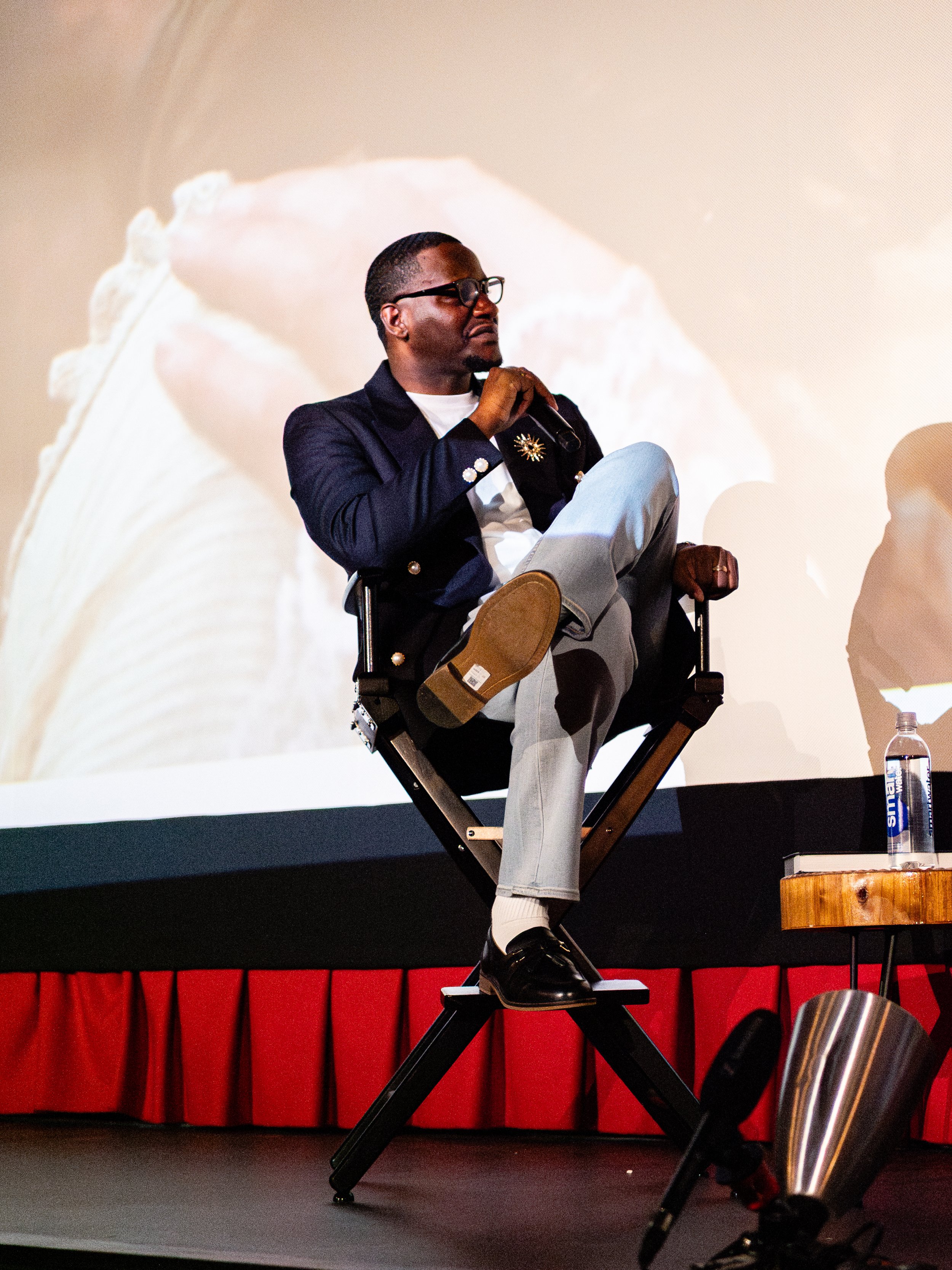 Man sitting on a stool with a backrest on stage, holding a microphone and looking thoughtfully to the side. He is wearing glasses, a blazer with decorative buttons, light-colored pants, and black shoes. There is a water bottle on a small round wooden