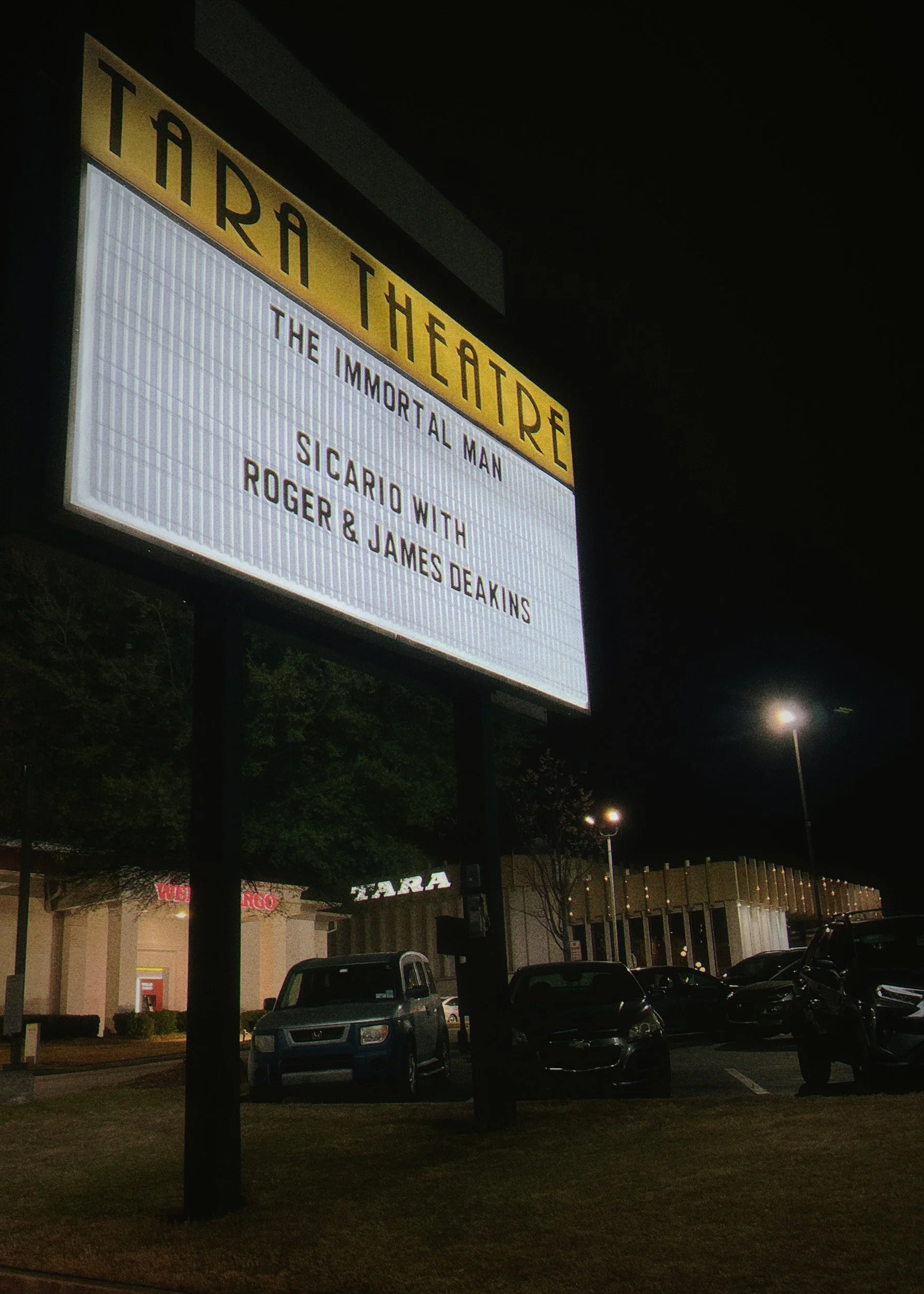 Nighttime scene of a shopping center parking lot with a lit marquee sign reading 'Theatre' and another sign indicating 'The Immortal Man' playing with Sicario with Roger & James Deakins, with parked cars and a lit building in the background.