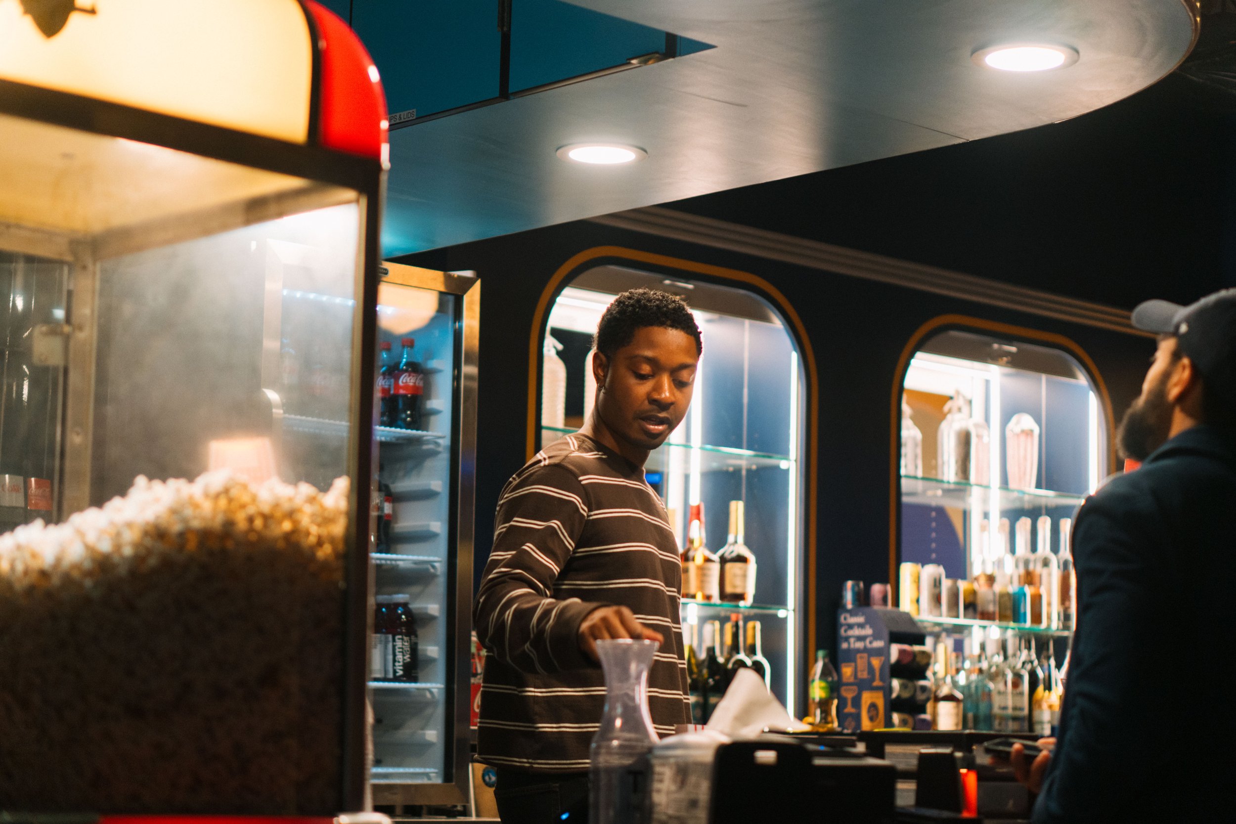 A man with dark curly hair wearing a striped shirt is working behind the counter in a cafe or bar, with another person in a cap partially visible on the right. The background includes illuminated shelves with bottles and a fridge with Coca-Cola, and 