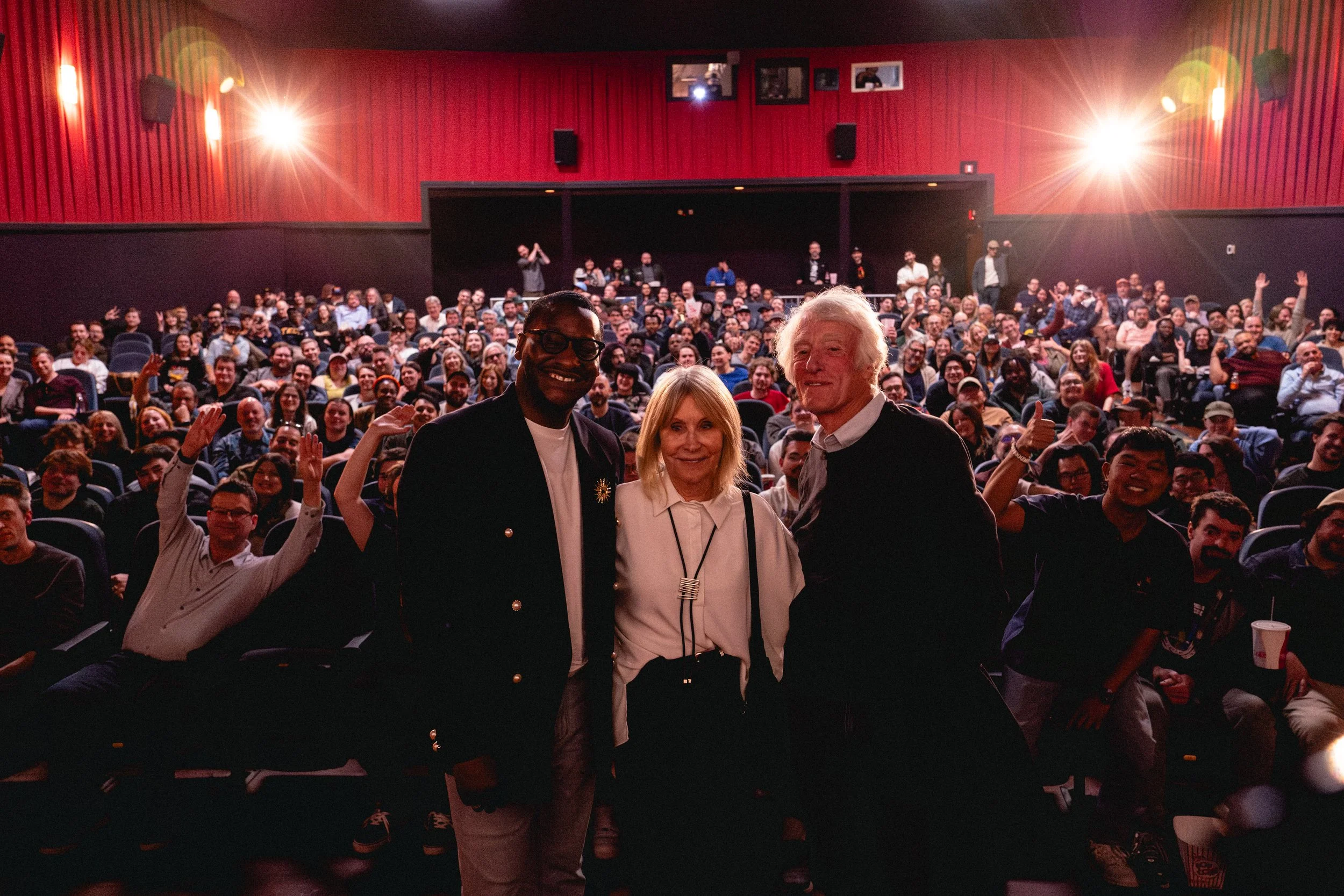 People in a theater with red walls taking a selfie in front of a large audience, some waving and giving thumbs up.