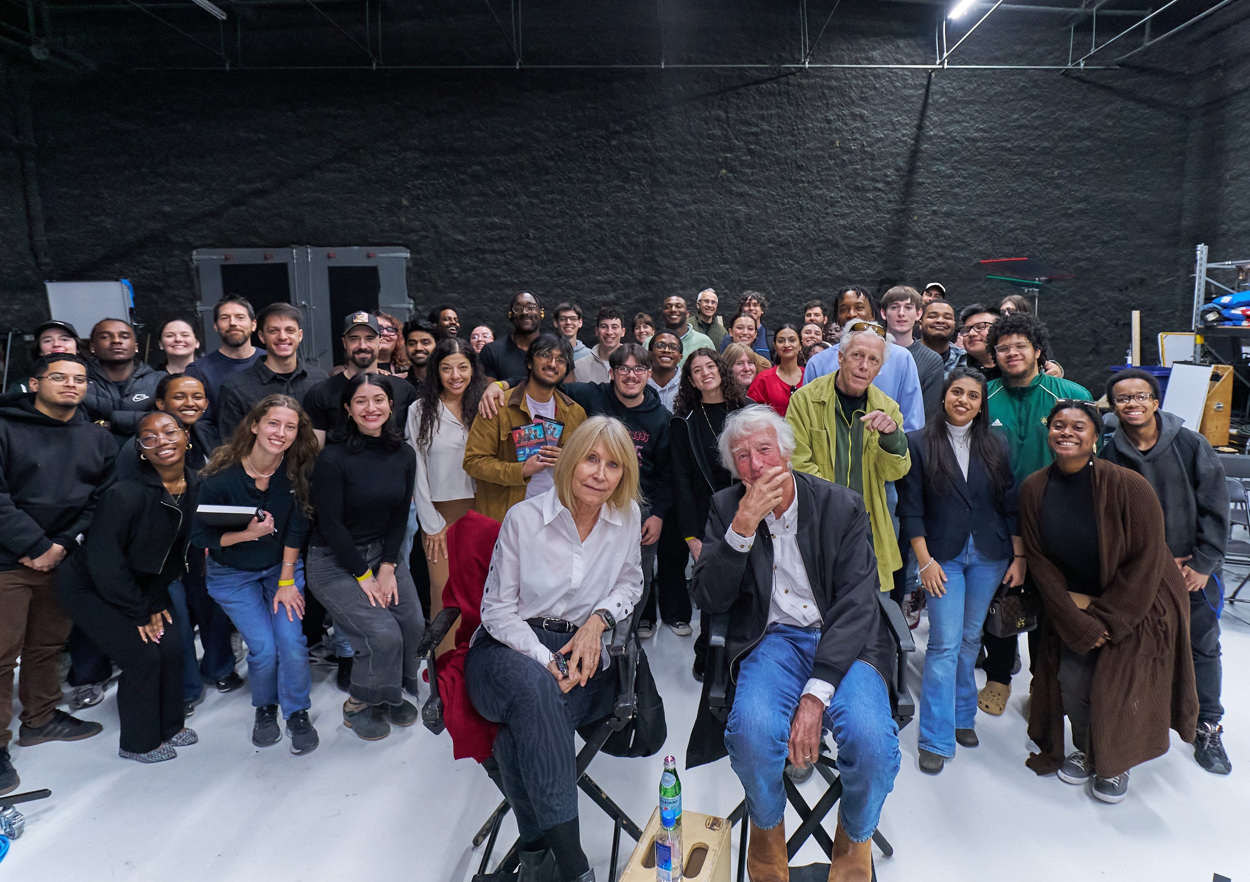 Group of diverse people gathered in an indoor space, smiling and posing for a group photo. Some are standing, while a few are sitting in the front, with a black textured wall in the background.