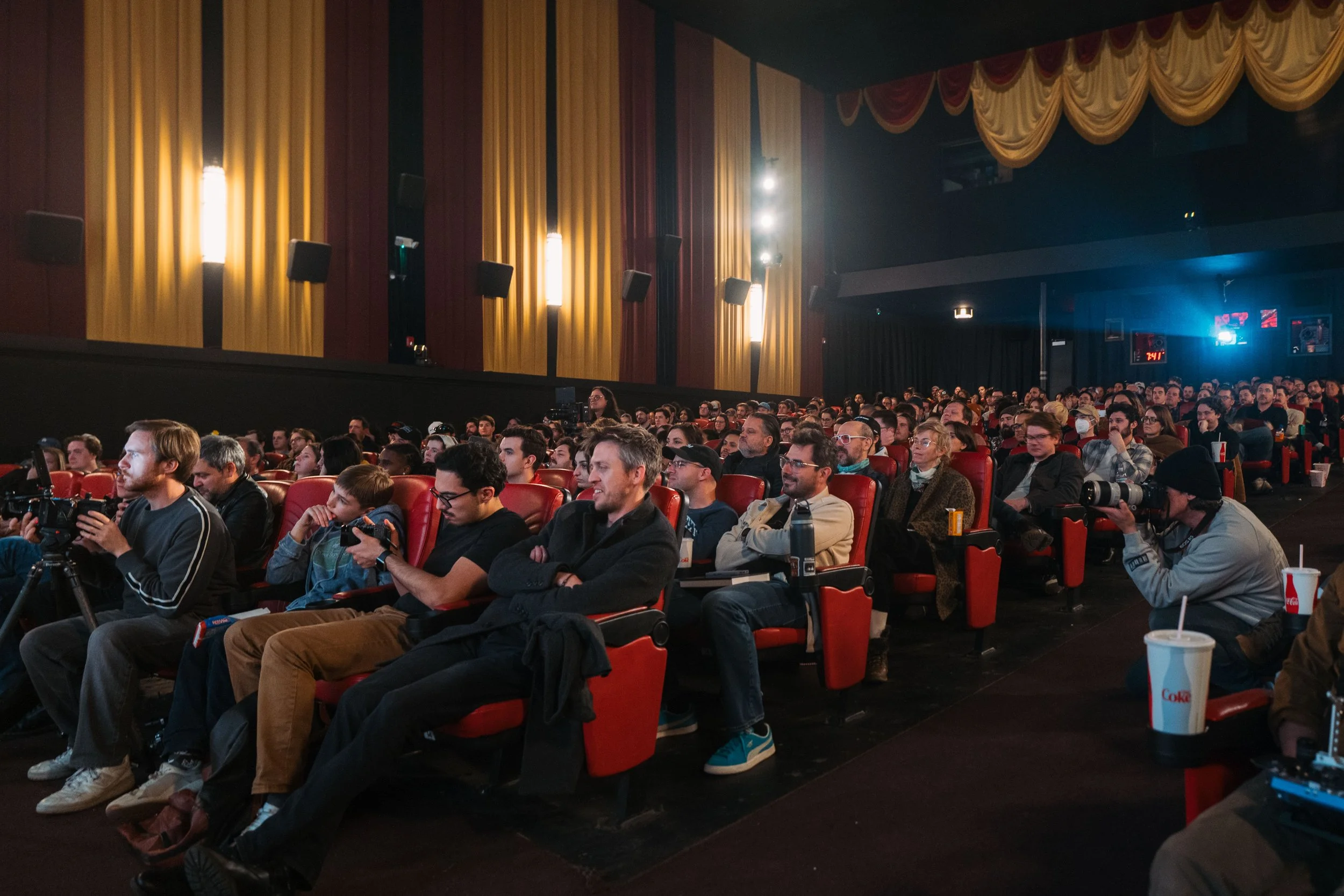 Audience seated in a theater, watching a presentation or movie, with some holding cameras or smartphones, and drinks on the armrest.